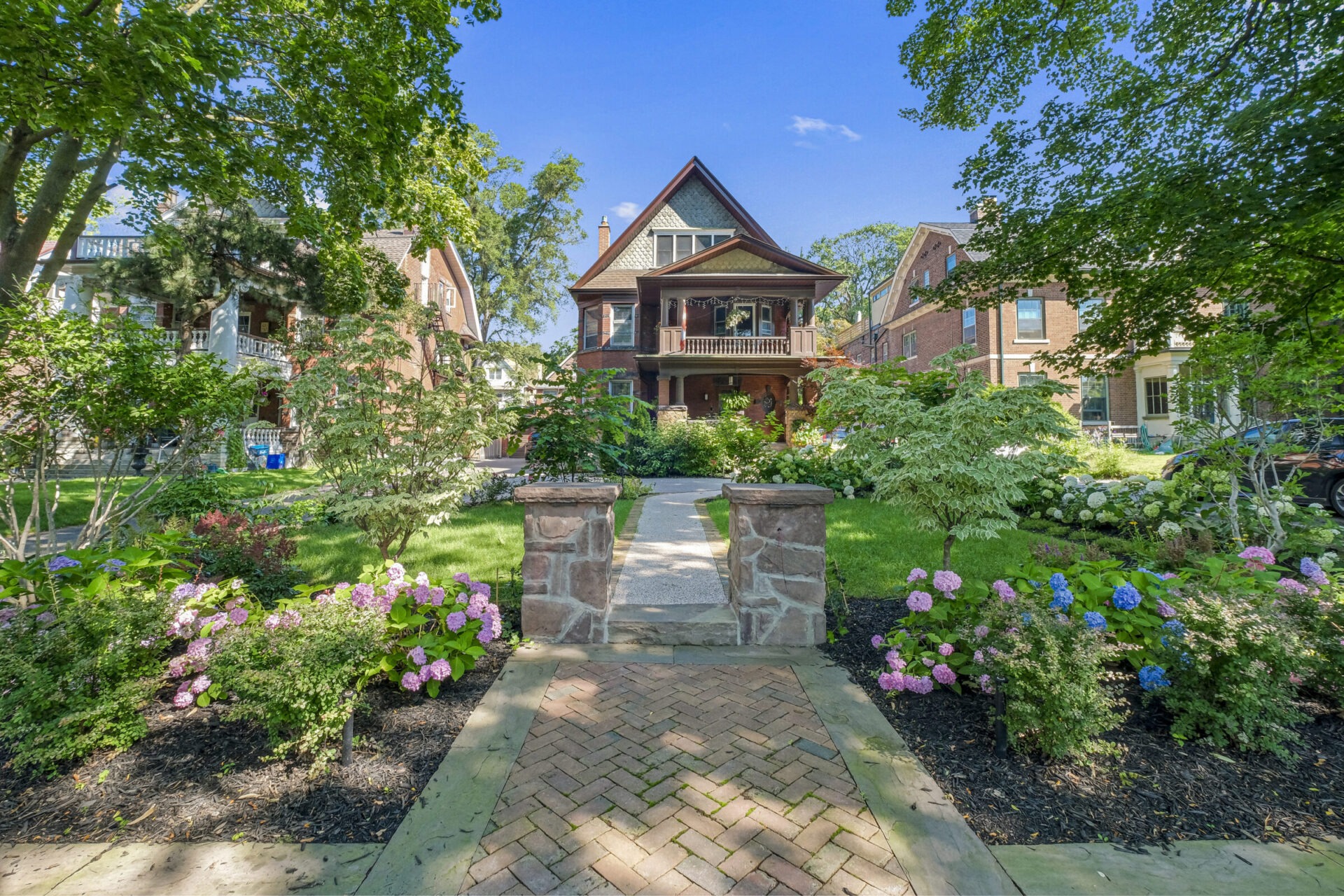 An elegant brick house with a peaked roof and front porch sits behind a landscaped garden with hydrangeas, under a blue sky with green foliage.