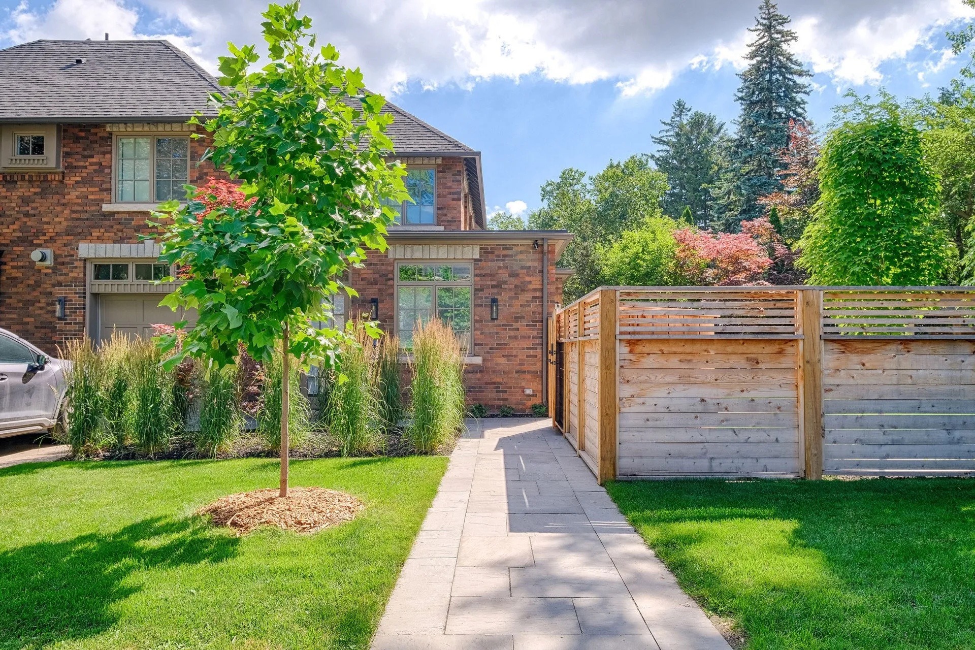 A suburban brick house with a lush green lawn and a young tree, bordered by a wooden fence and a stone pathway under a clear sky.