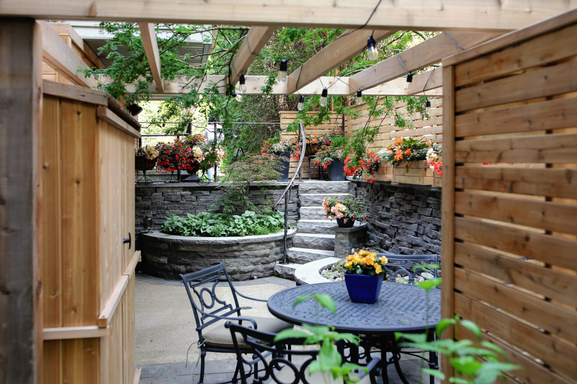 A serene outdoor patio area with wooden privacy walls, stone planters, lush greenery, hanging lights, a metal table, and chairs, creating a cozy atmosphere.