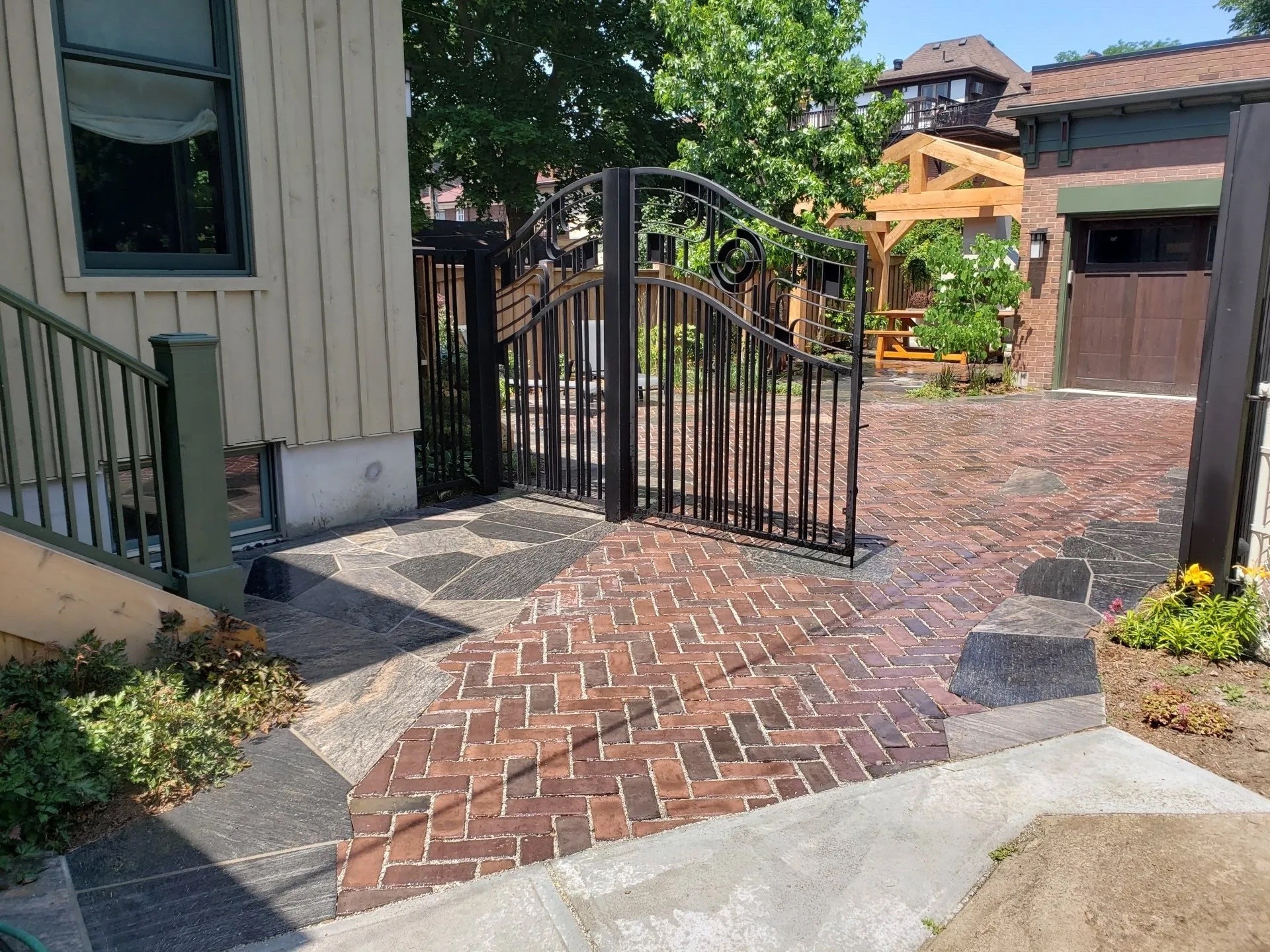 A residential outdoor area with a brick driveway, an open black gate, a wooden pergola, and greenery under a bright sky. No people are visible.