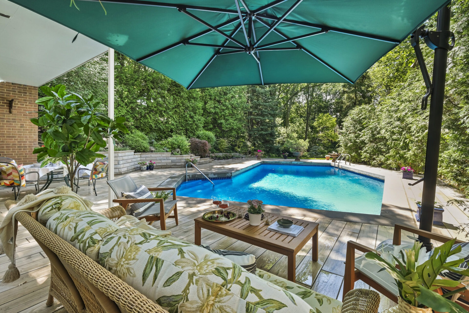 An inviting backyard with a large swimming pool, shaded patio area, comfortable outdoor furniture, lush greenery, and a clear blue sky above.