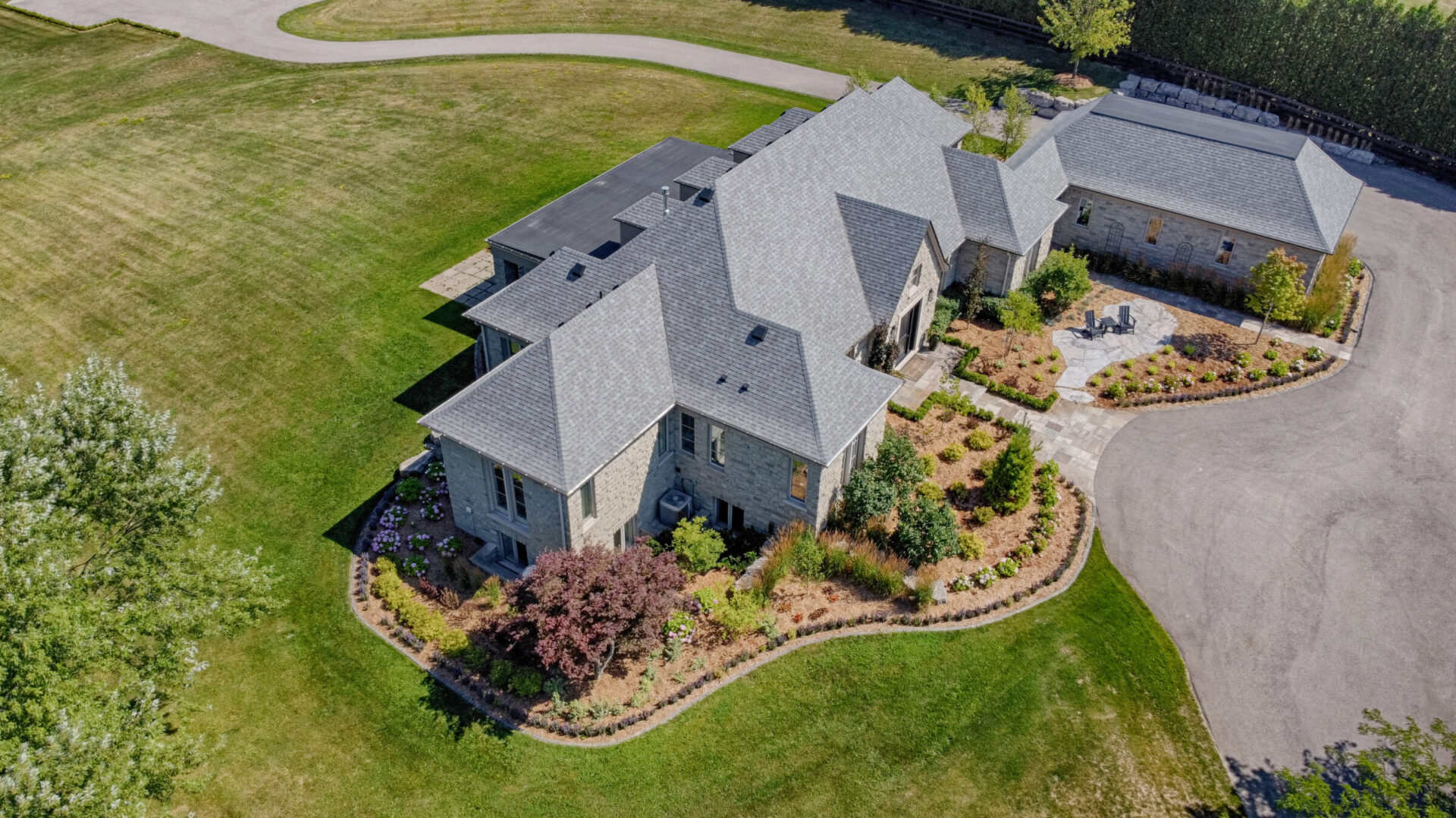 An aerial view of a large stone house with a grey roof, surrounded by manicured lawns, a circular driveway, and landscaped gardens.