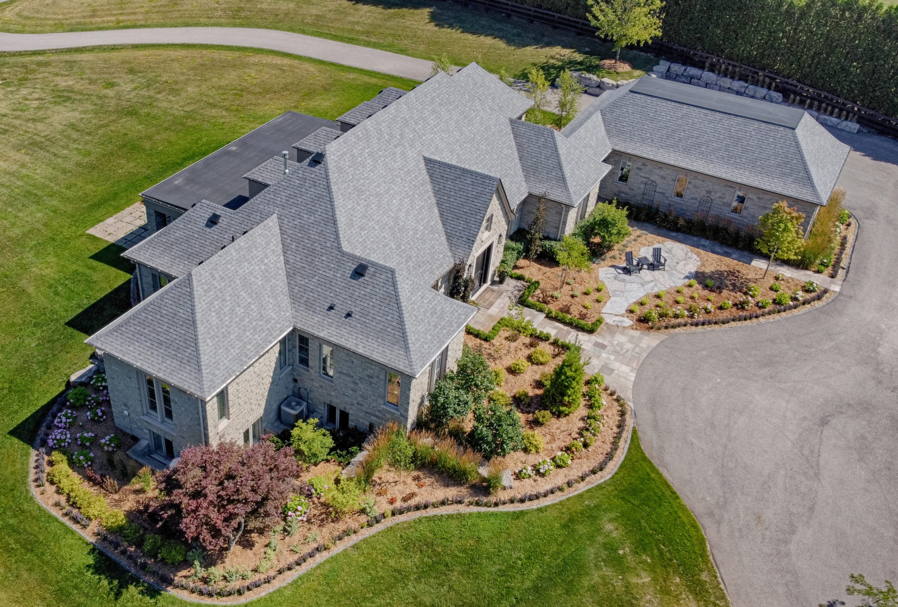 An aerial view of a large stone house with a grey roof, surrounded by manicured lawns, a circular driveway, and landscaped gardens.