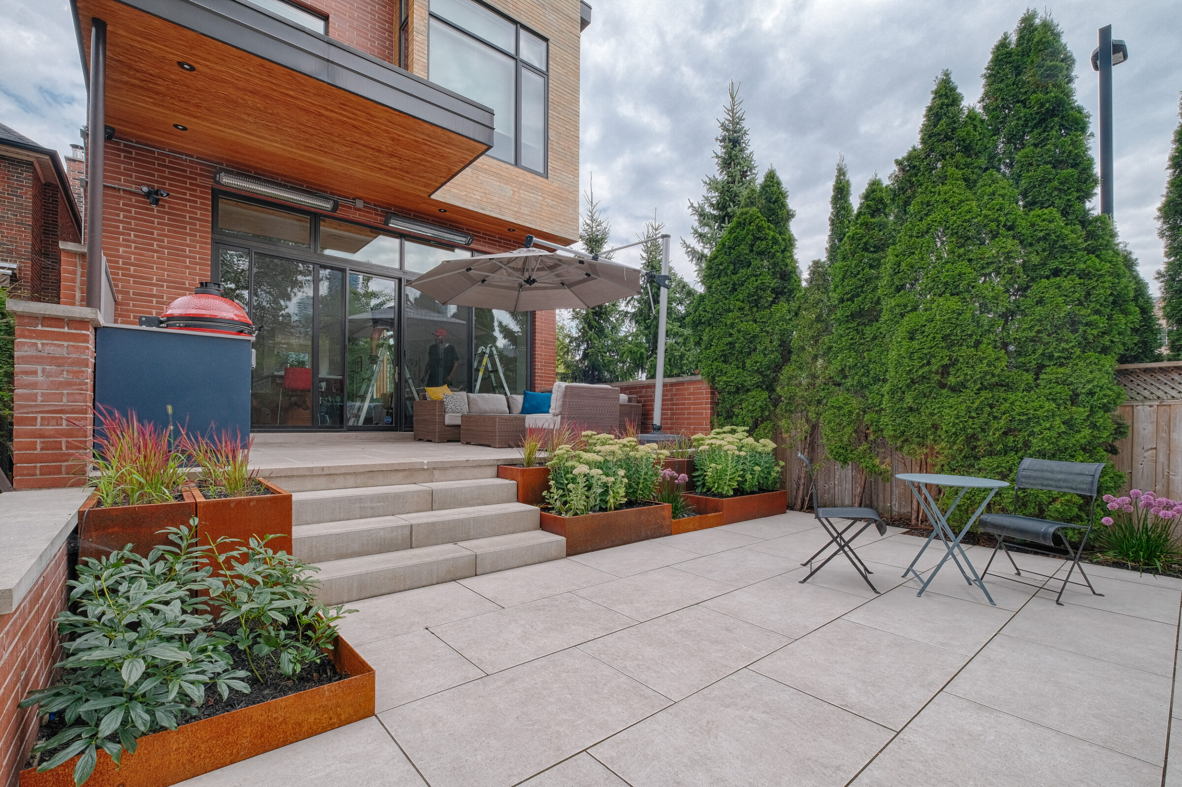A spacious patio with modern furniture, a grill, and lush greenery beside a contemporary brick house with large glass doors and wooden accents.
