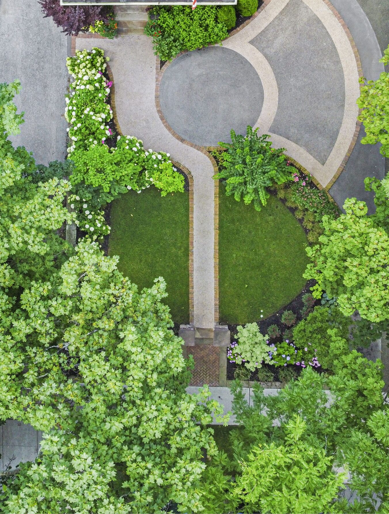 Aerial view of a manicured garden with winding pathways, lush greenery, flowering shrubs next to a section of a residential building roof.
