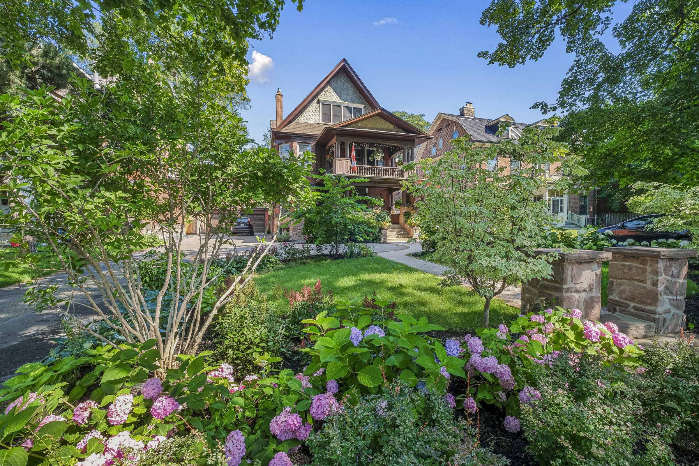 An elegant house with a gabled roof and wooden balcony, surrounded by lush greenery and blooming hydrangeas in a peaceful residential setting.