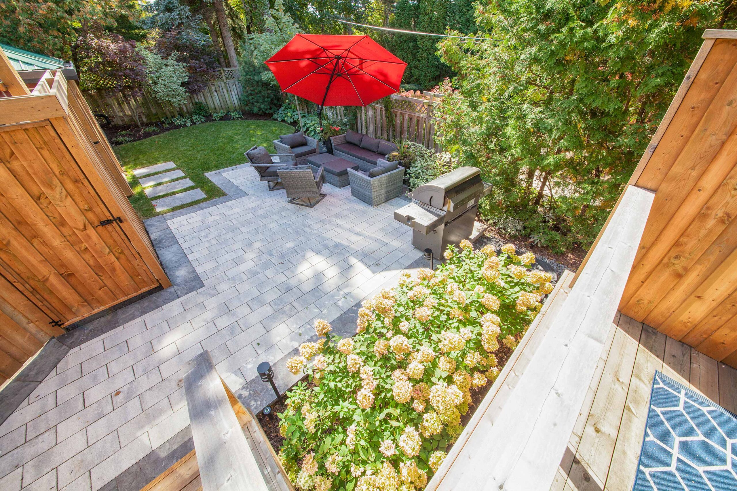 This image shows a well-manicured backyard with a patio area, outdoor furniture, a red umbrella, a barbecue grill, and lush greenery surrounding the space.