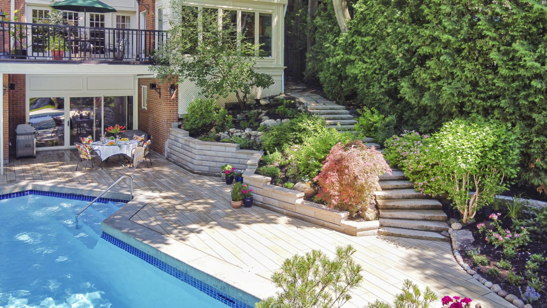 A landscaped backyard with a swimming pool, stone steps, lush greenery, a dining setup under a balcony, and a clear sunny sky.