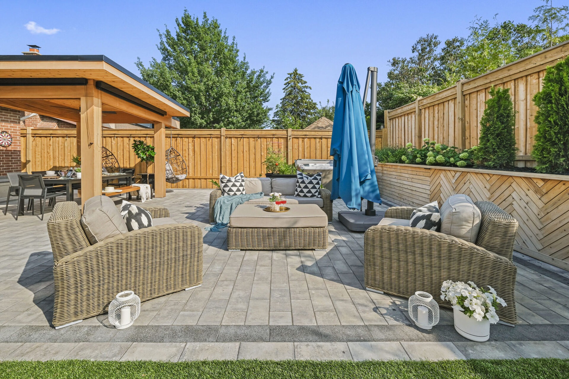 An inviting backyard patio with wicker furniture, a pergola, lush greenery, a closed blue umbrella, and decorative cushions under a clear sky.