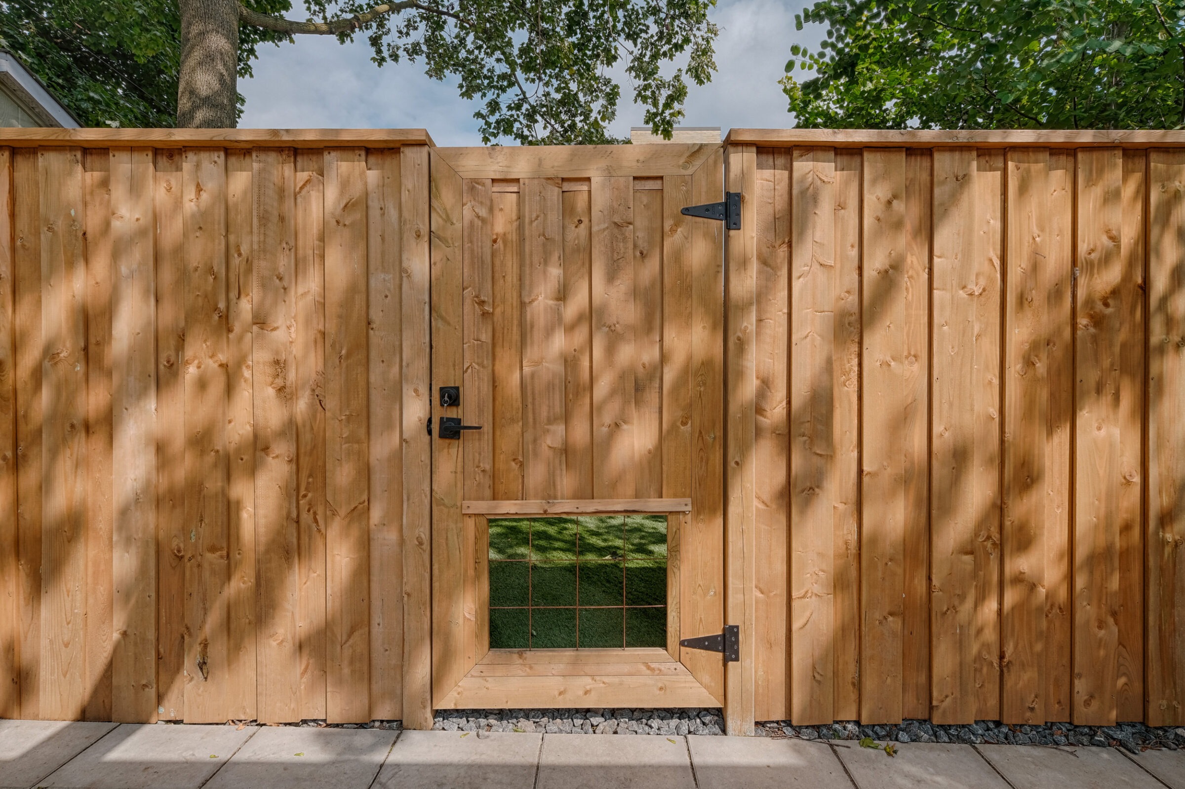 This image showcases a tall wooden fence with a closed gate. The gate has black hardware, a small window with a grid design, and is sunlit.