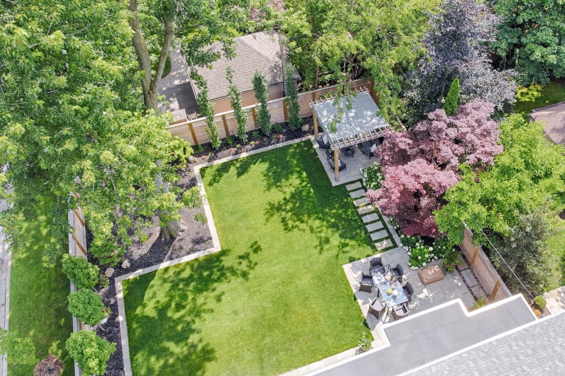 An aerial view shows a well-manicured backyard with lush grass, a pergola, patio furniture, diverse plants, and trees casting shadows.