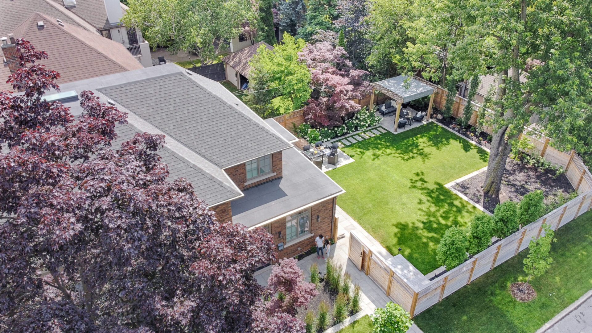 Aerial view of a suburban landscape showing two houses with shingled roofs, a manicured lawn, trees in blossom, and a wooden fence surrounding the property.