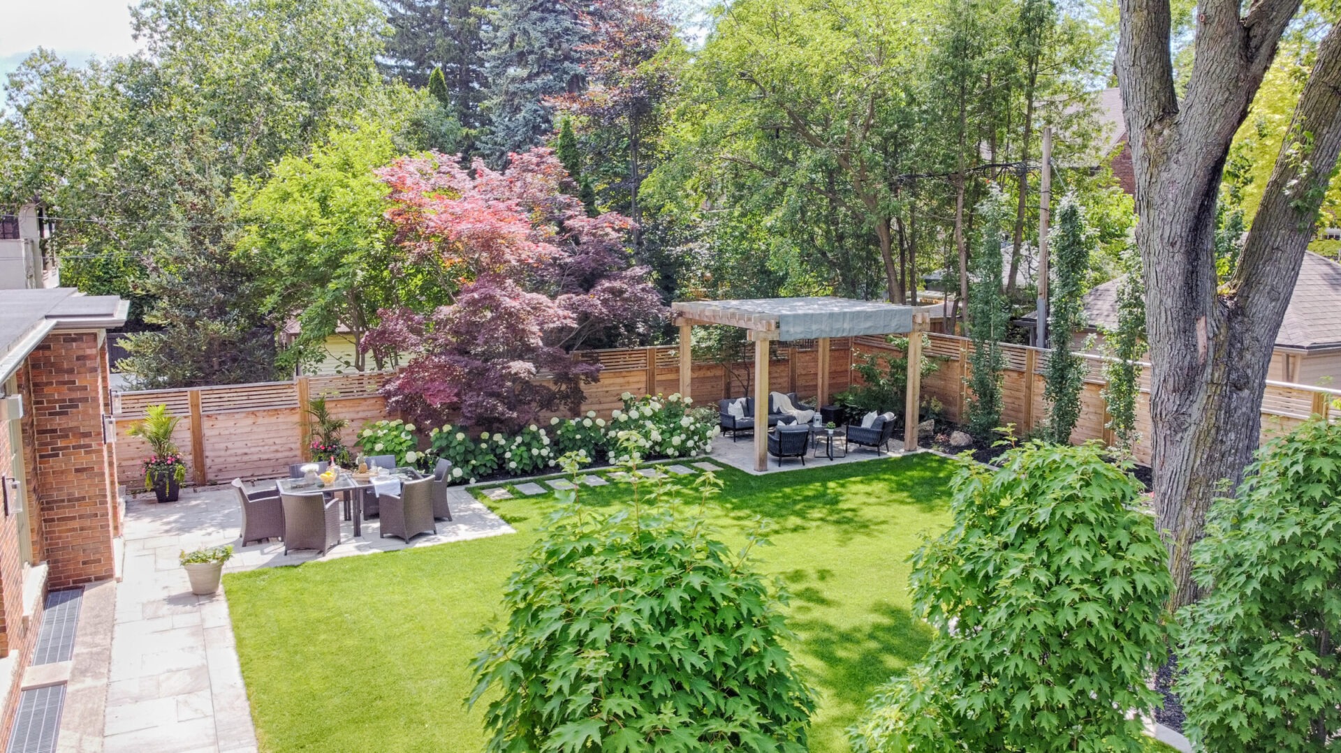 An aerial view of a lush backyard with green lawn, a pergola, outdoor seating, trees, and a wooden fence in a residential area.