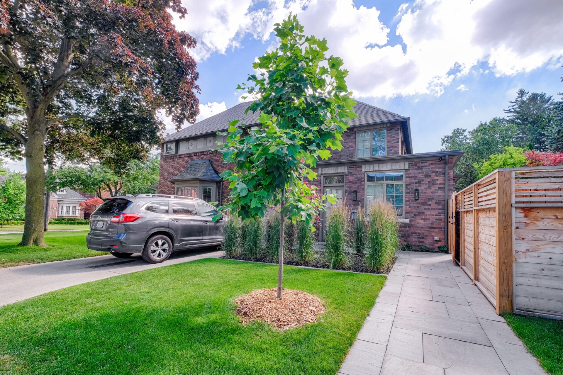 A brick house with a dark roof features a driveway on the left where a grey SUV is parked. Greenery surrounds the home including a young tree.