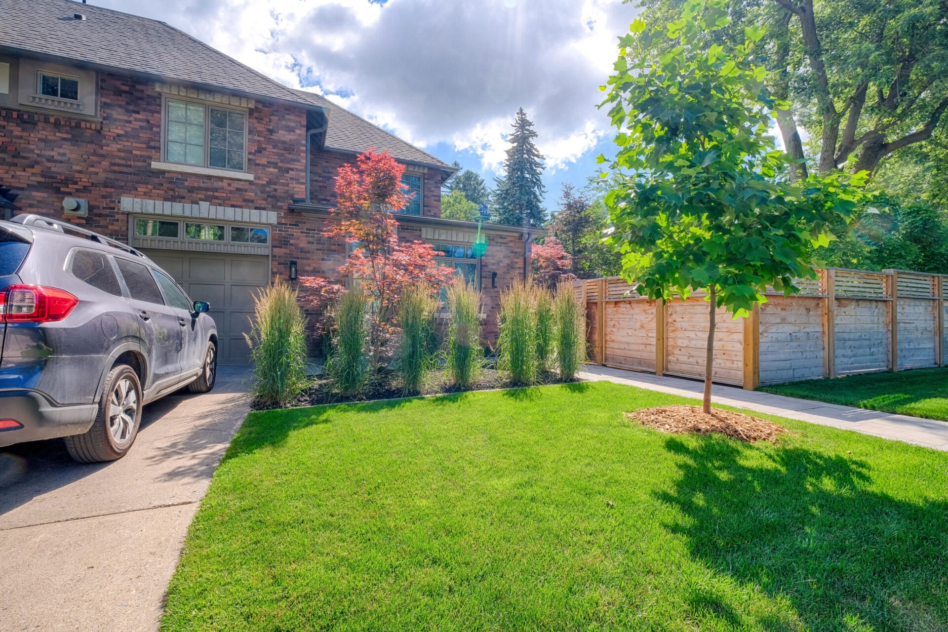 A brick house with a garage, a parked SUV, a well-maintained lawn, ornamental plants, a wooden fence, and a young tree under a blue sky.