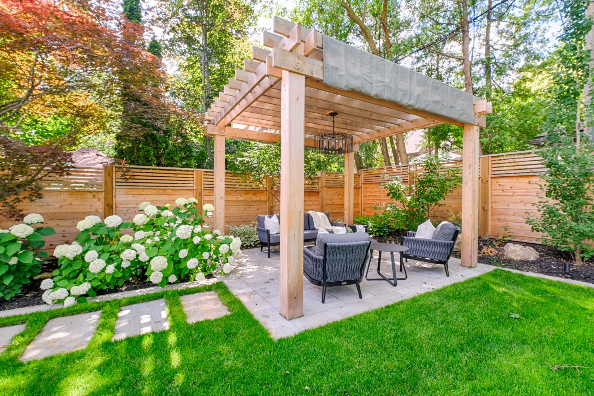 A serene garden with a wooden pergola, comfortable outdoor furniture, lush green lawn, hydrangea bushes, and a privacy fence under a clear sky.