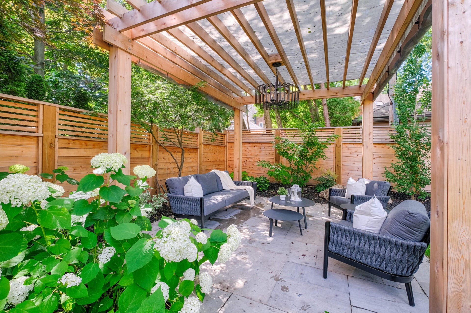 An inviting outdoor patio area with comfortable furniture under a wooden pergola, surrounded by lush greenery and blooming white hydrangeas.