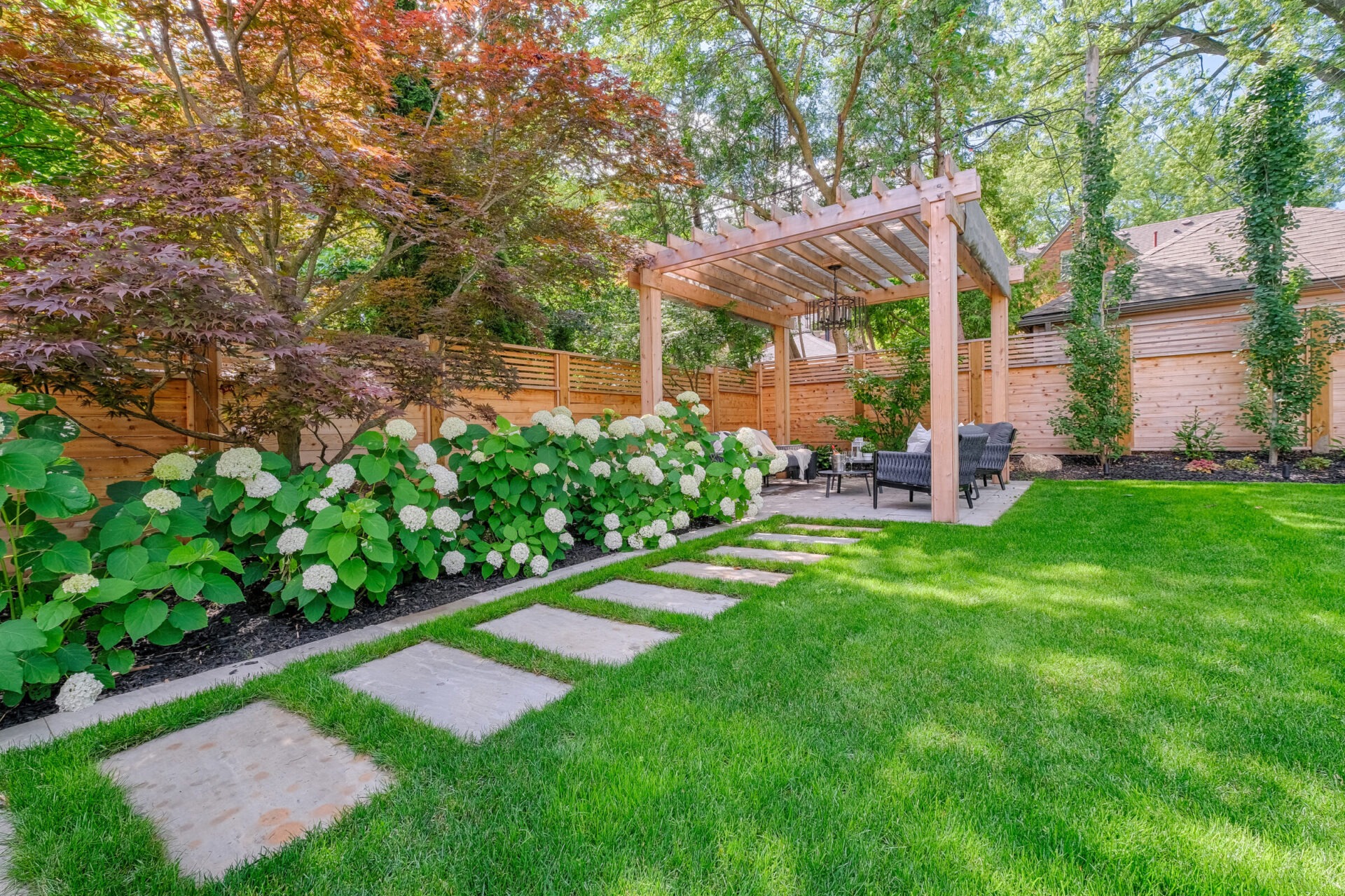 This image shows a well-manicured garden with stepping stones, a lush lawn, flowering shrubs, a wooden pergola, and comfortable outdoor furniture in a serene setting.