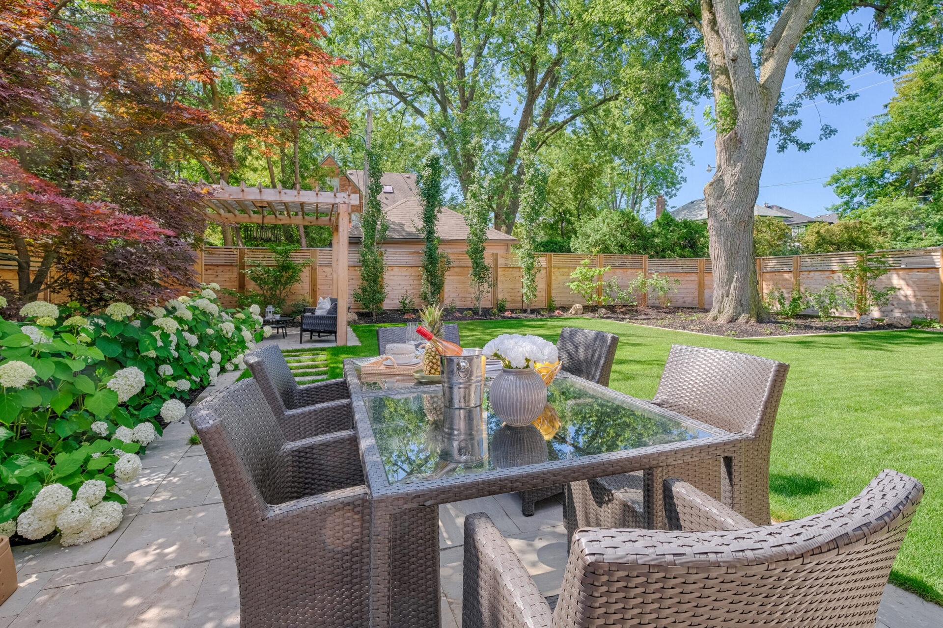 A vibrant backyard with lush green grass, colorful hydrangeas, mature trees, a pergola, and a wicker patio dining set with a glass-topped table.