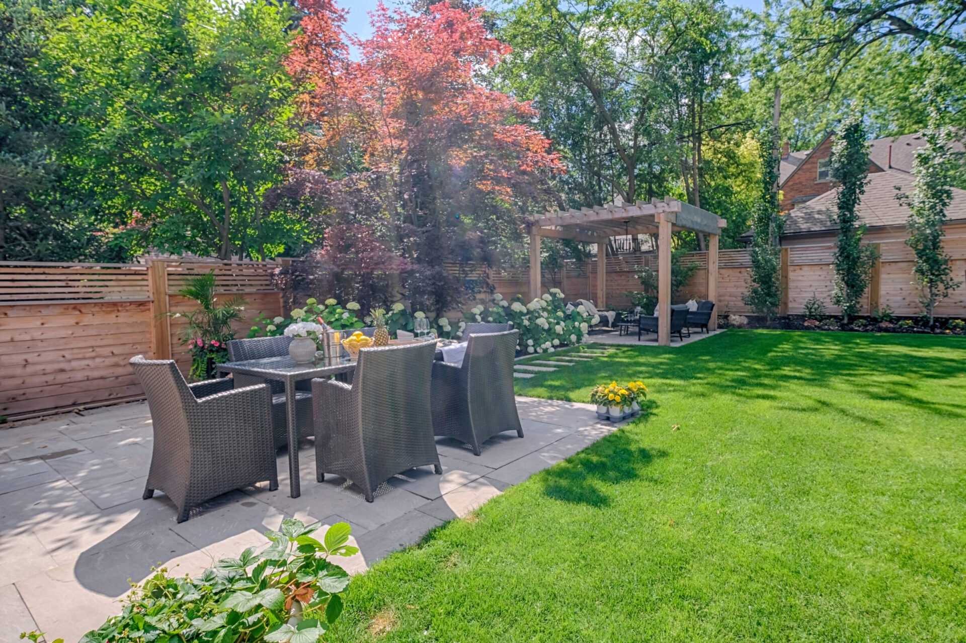 This is a serene garden with a dining set, lush lawn, vibrant plants, a pergola, and a wooden fence under a clear blue sky.