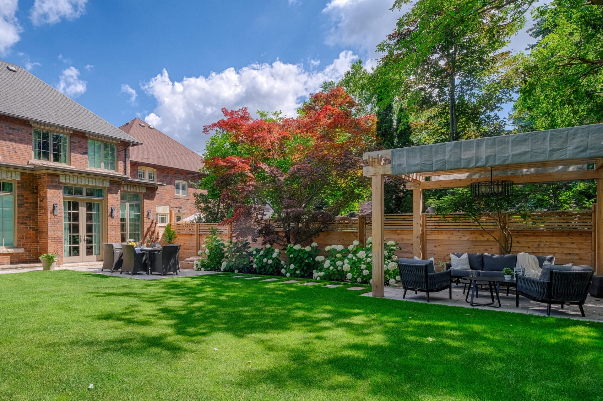 A lush backyard with manicured lawn, cozy outdoor seating, colorful foliage, under a clear sky beside a brick house with large windows.