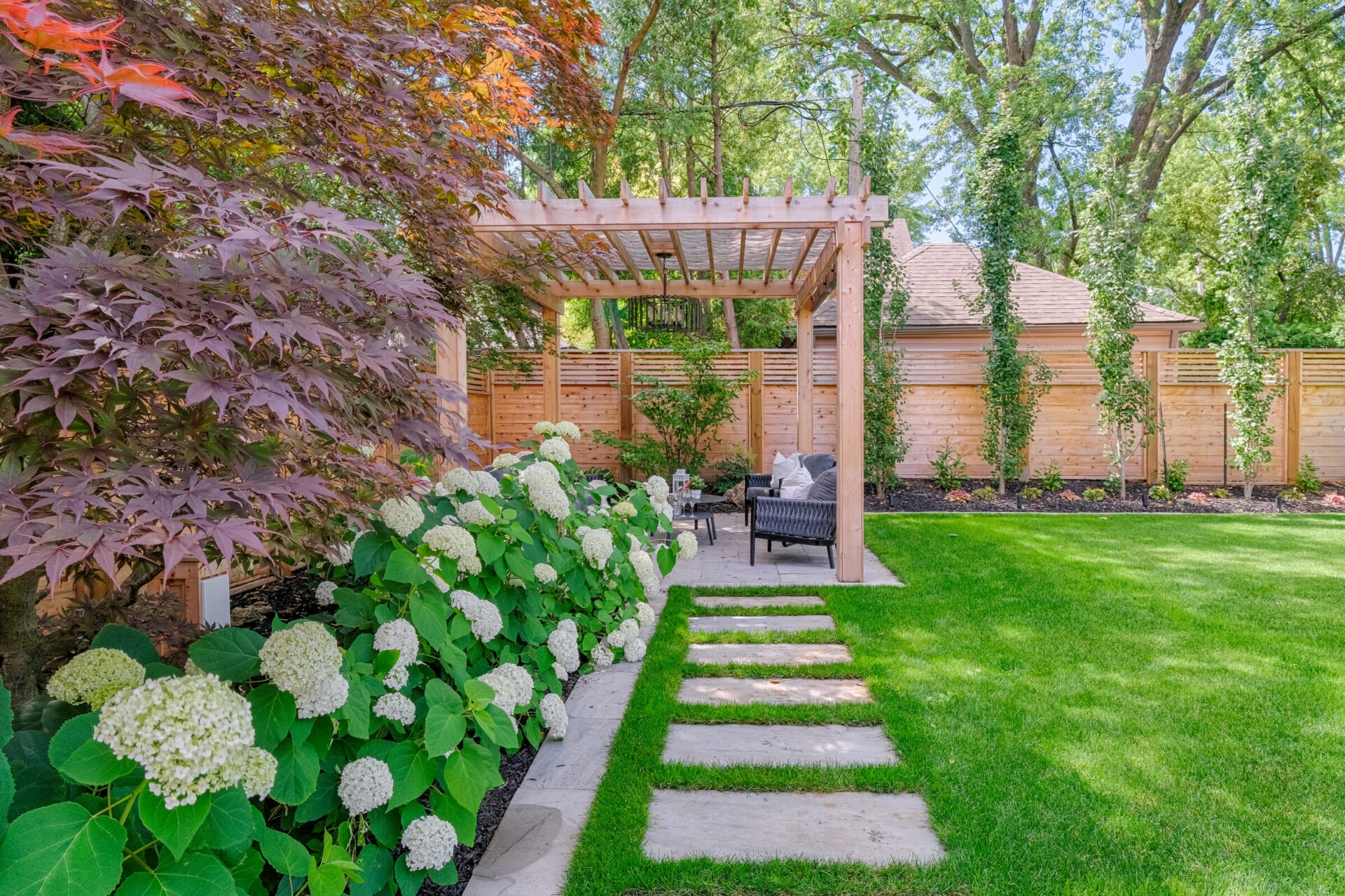 This image shows a beautiful garden with vibrant green grass, flowering shrubs, stepping stones, a pergola, and outdoor seating surrounded by wooden fences.