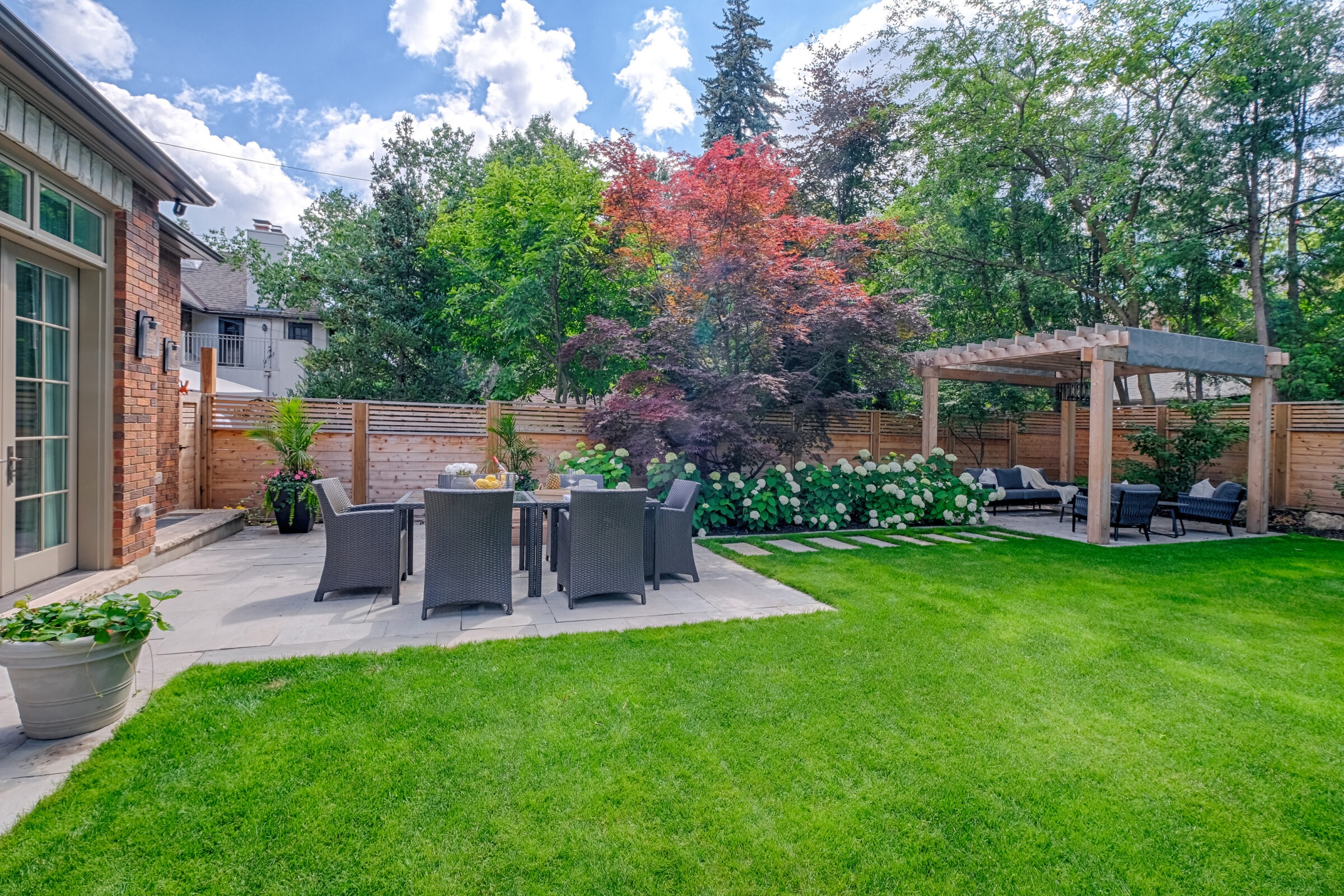 A well-manicured garden with lush green grass, a dining area, a pergola, various trees, a wooden fence, and a brick building on a sunny day.