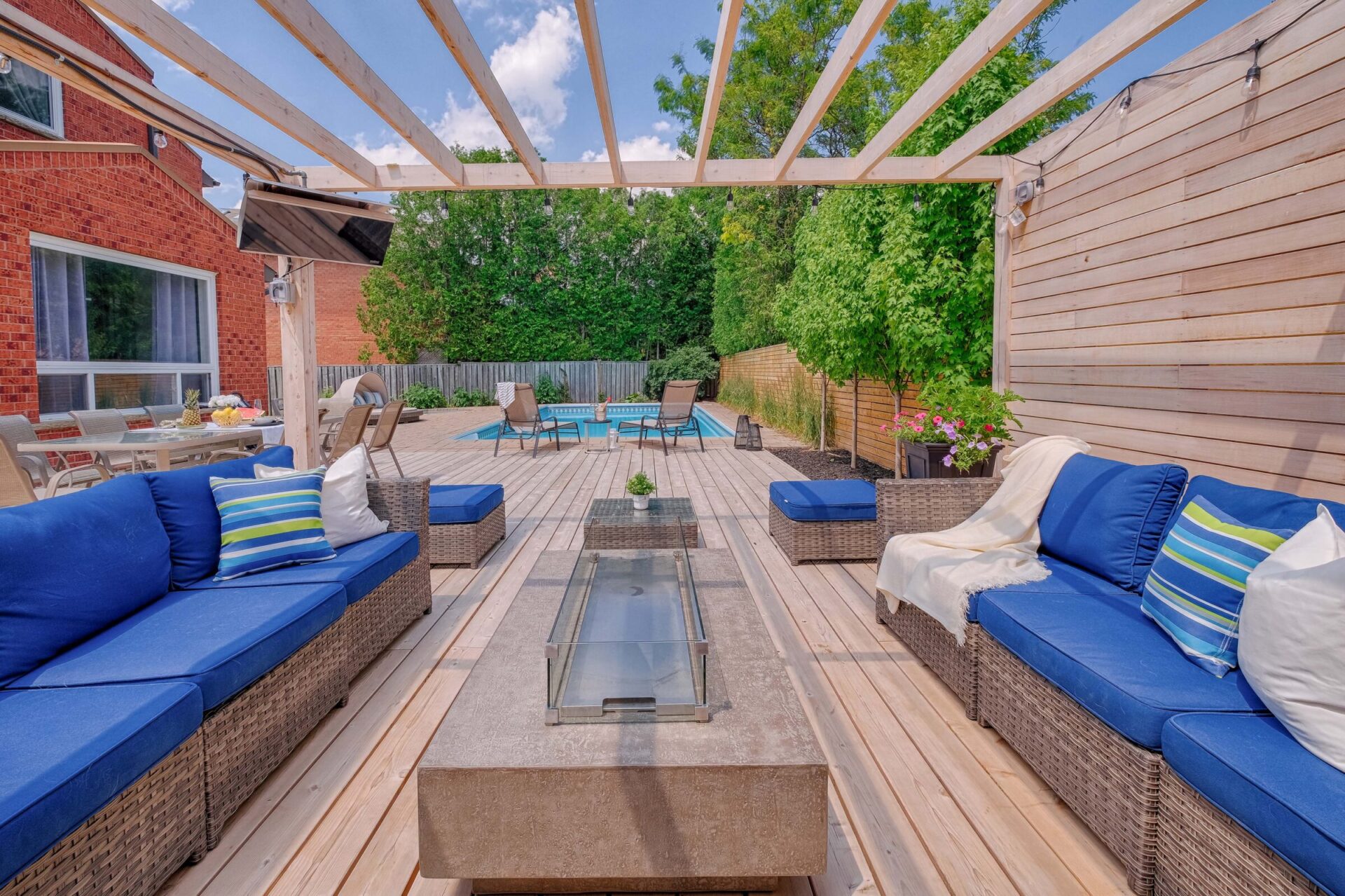 An outdoor patio with a pergola, blue cushioned seating, glass table, and a pool in the background. Sunny sky, lush green trees, and wooden decking.