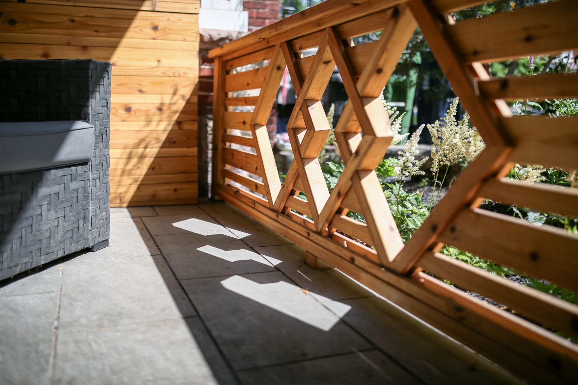 A wooden patio with geometric railing patterns casting shadows on tiled flooring, alongside outdoor wicker furniture and garden greenery peeking through.