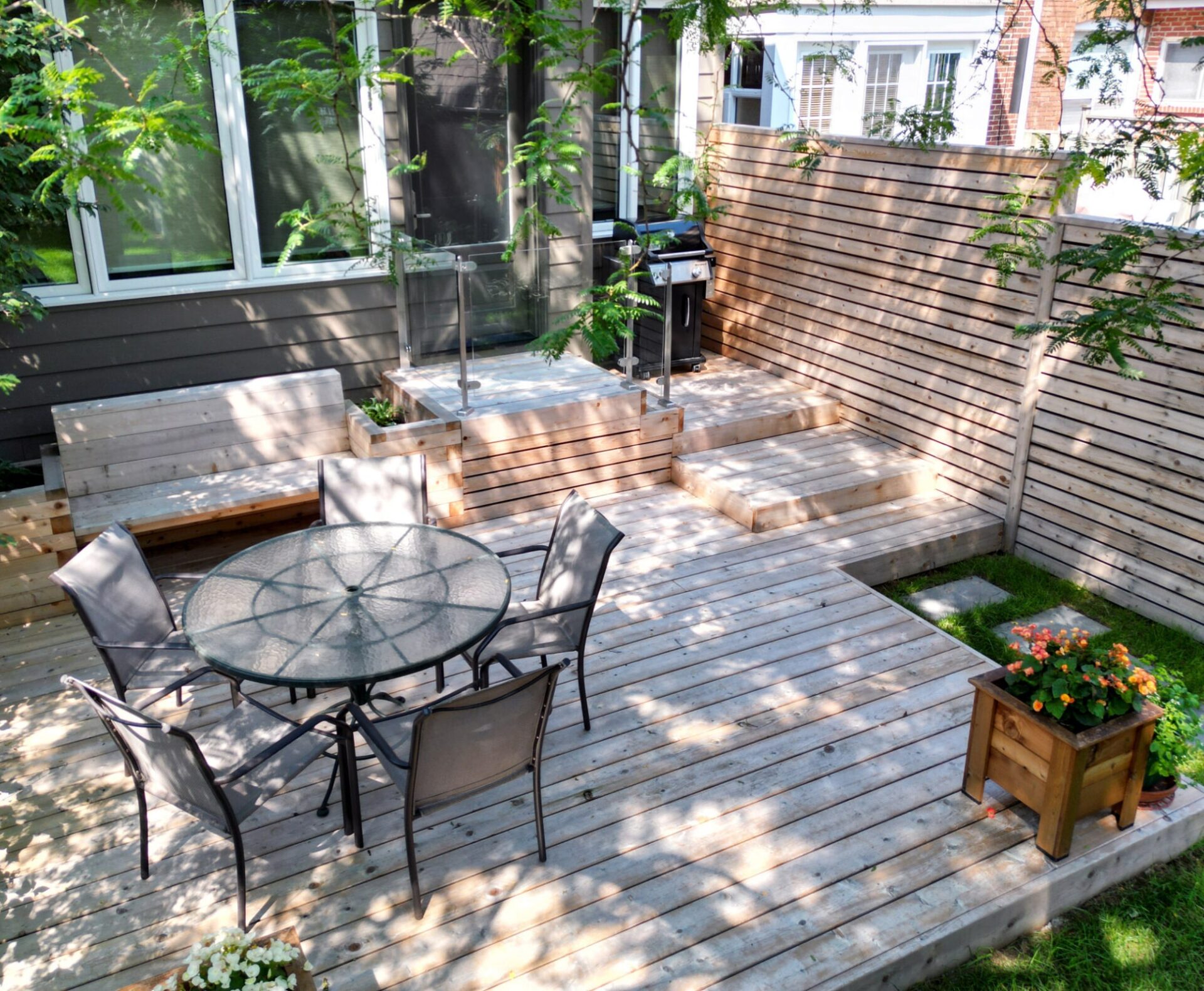 An outdoor wooden deck with steps and a dining set, adjacent to a house, surrounded by a slatted fence, plants, and a barbecue grill.