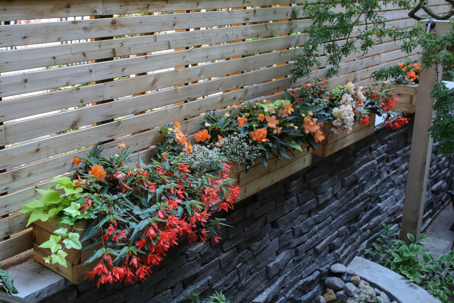 A wooden fence with integrated flower boxes filled with blooming flowers over a dark gray stone wall and a pebbled ground beneath.