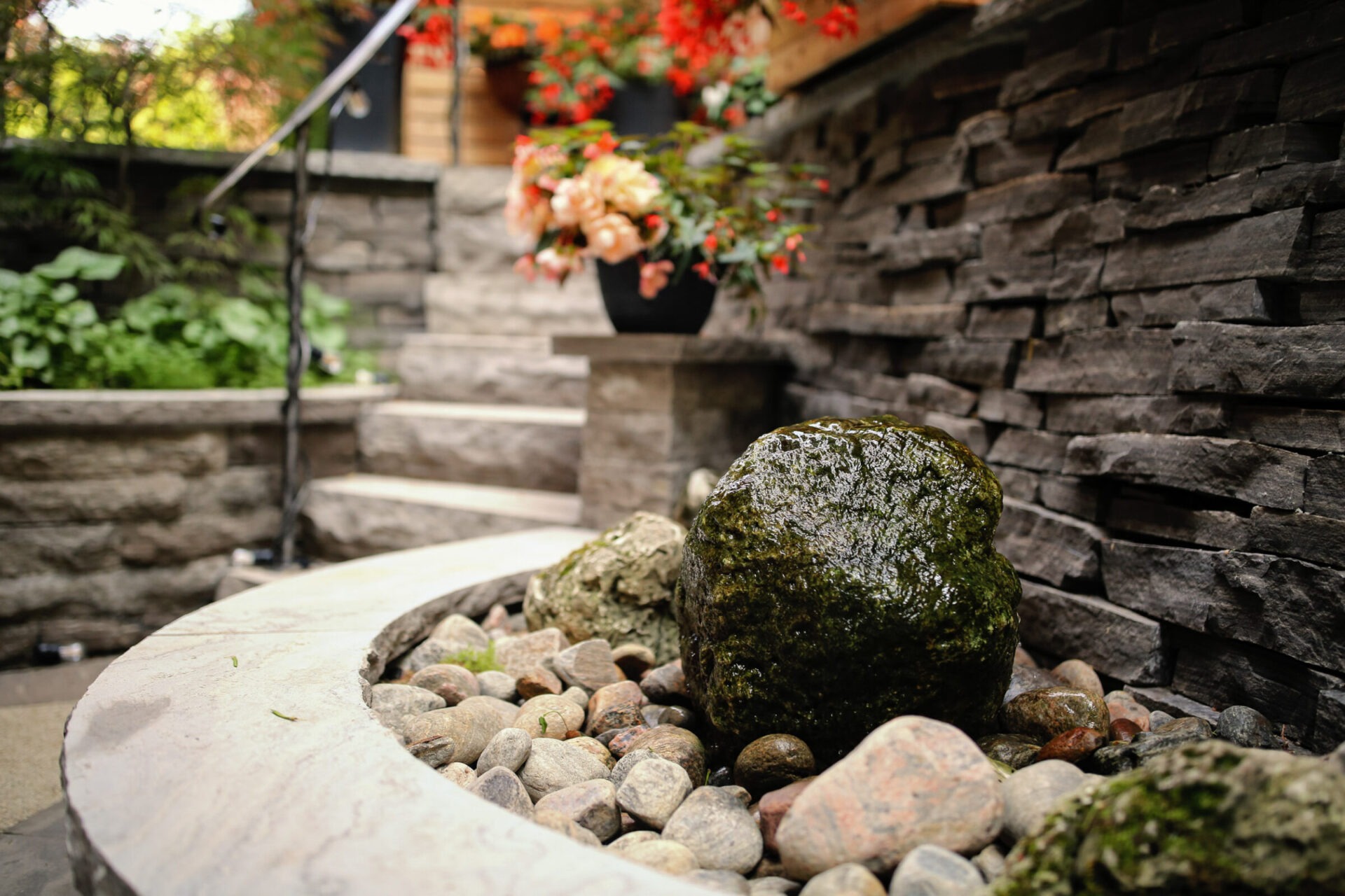 An outdoor scene with a moss-covered rock in a pebble-filled basin, stone steps, a metal railing, and potted flowers against a brick wall.