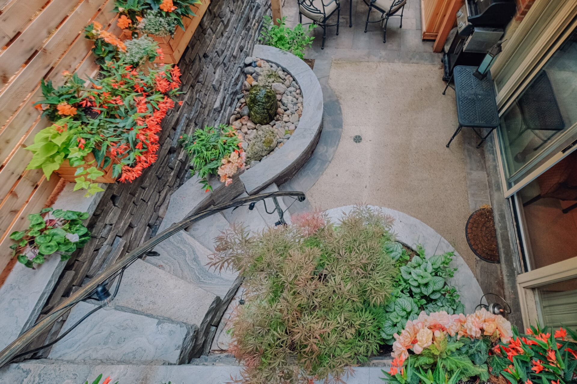 An outdoor space with stone stairs surrounded by vibrant flowers and plants, featuring a curved retaining wall and patio furniture.