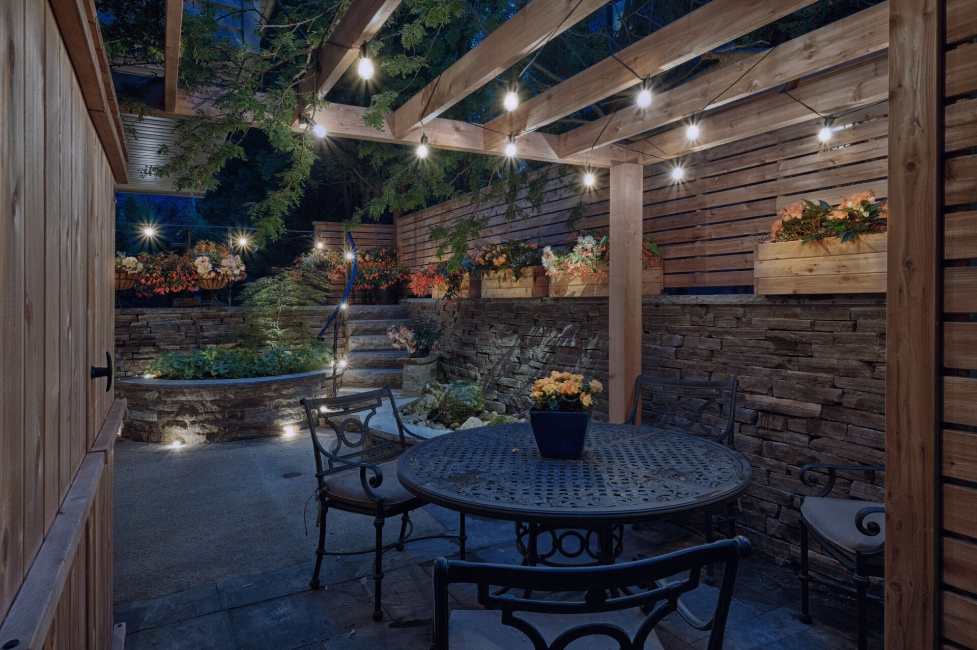 An outdoor patio area at dusk with lighting, wooden walls, stone stairs, a metal table, chairs, and decorative plants creating a cozy ambiance.