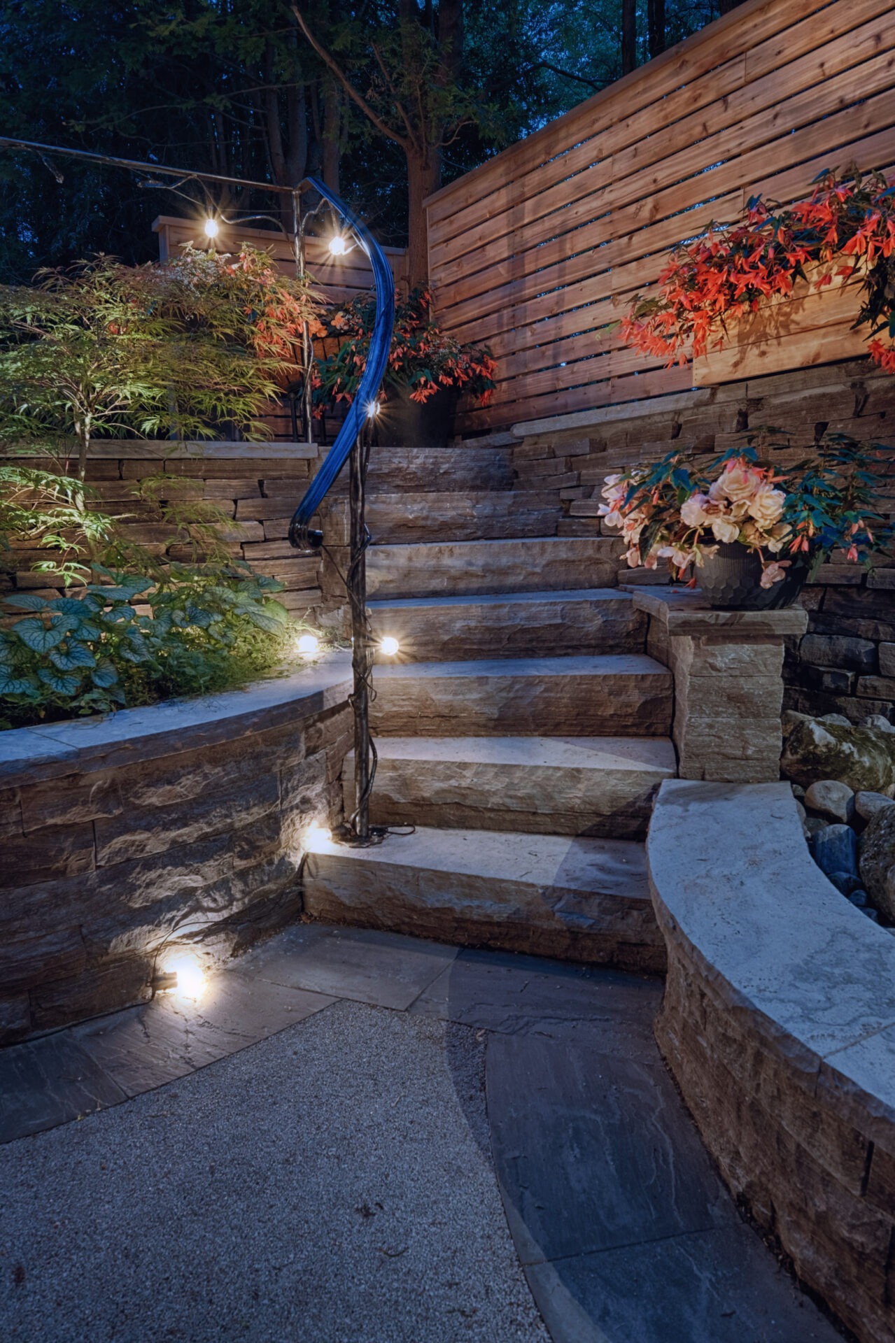 An outdoor stone staircase illuminated by built-in lights with wooden fencing, lush greenery, and potted flowers. It exudes a warm, inviting ambiance at dusk.