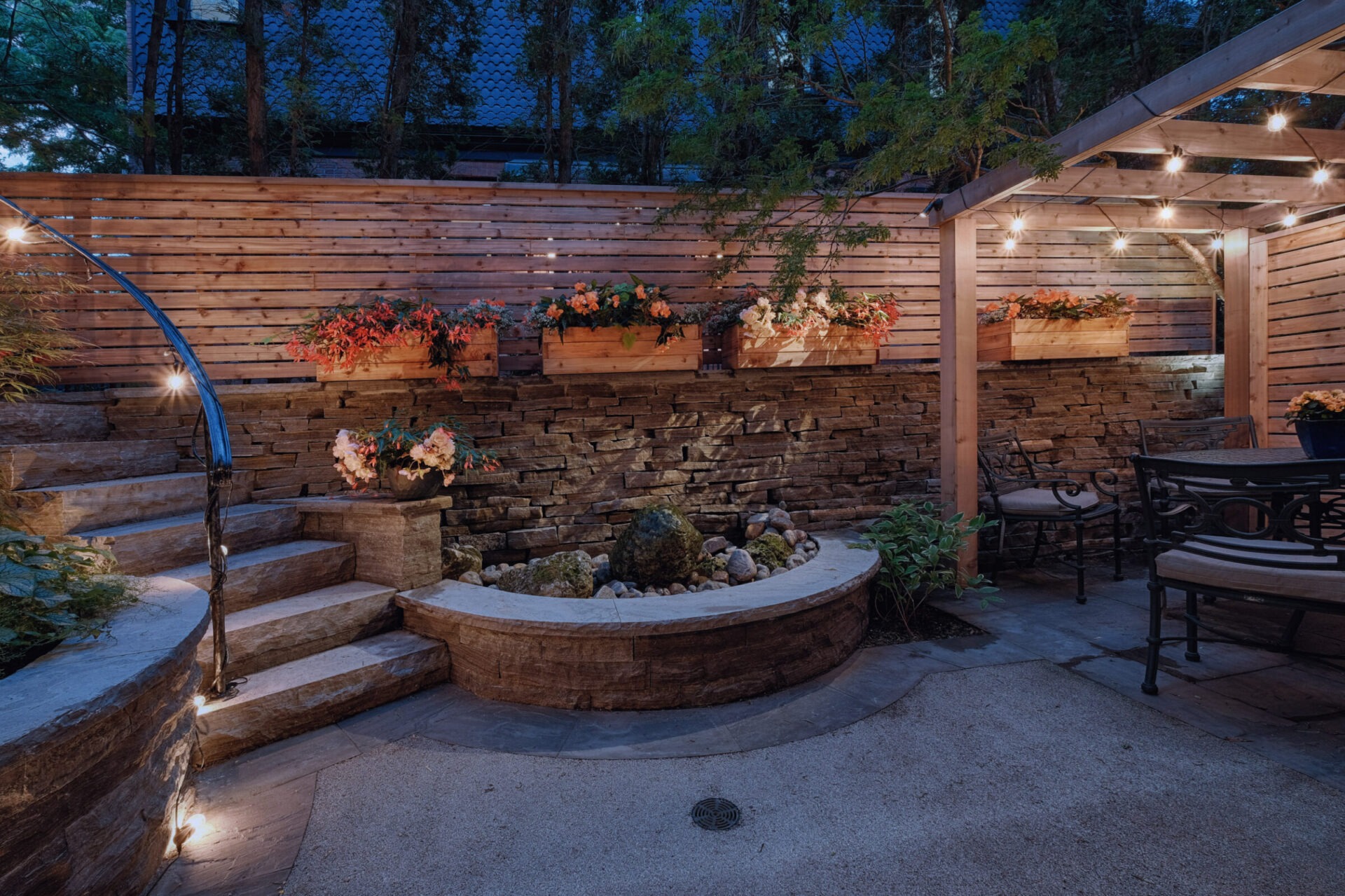 An inviting outdoor patio area with wooden benches, stairs, planter boxes, a water feature, and soft lighting under a pergola at twilight.