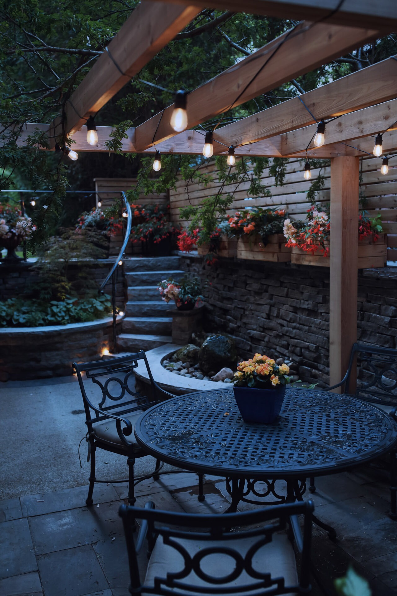 A cozy outdoor patio at dusk, featuring string lights, a wrought-iron table and chairs, steps leading to a garden with lush, flowering plants.