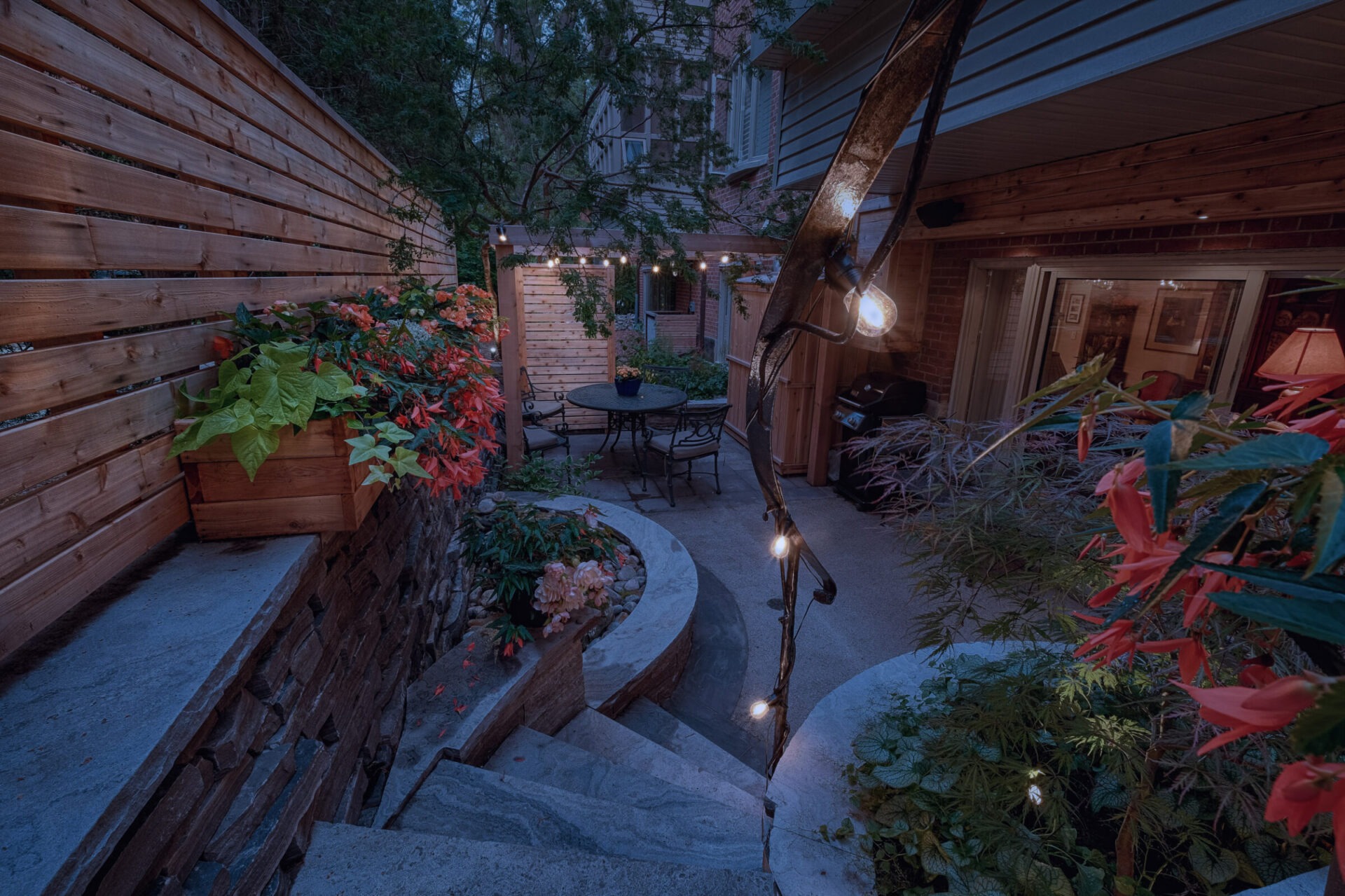 A tranquil outdoor patio area illuminated by string lights with lush plants, stone steps, and a sitting area with a table and chairs.