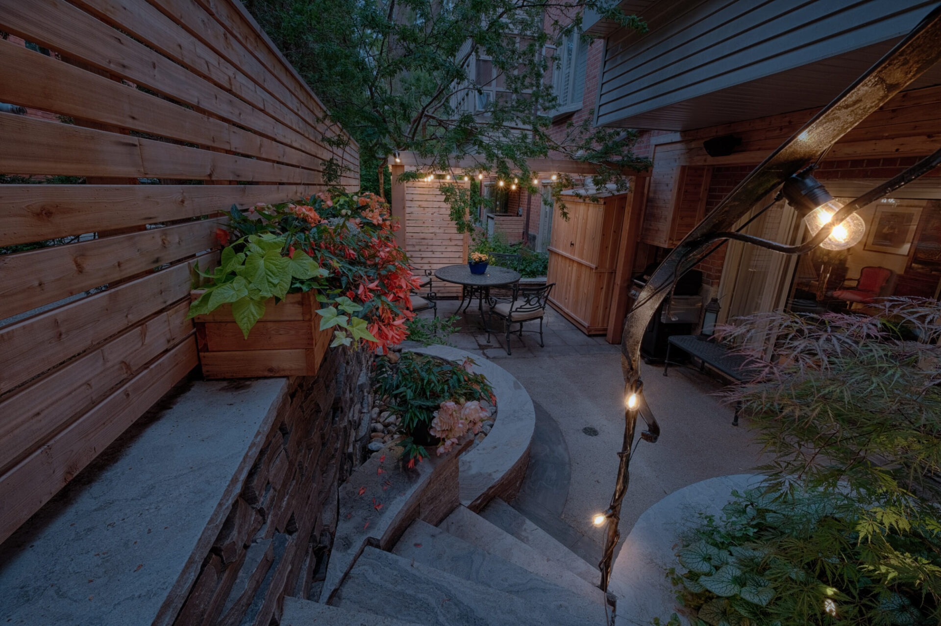 A cozy outdoor patio at dusk with string lights, a stone pathway, a seating area, lush plants, and a wooden fence in a residential area.