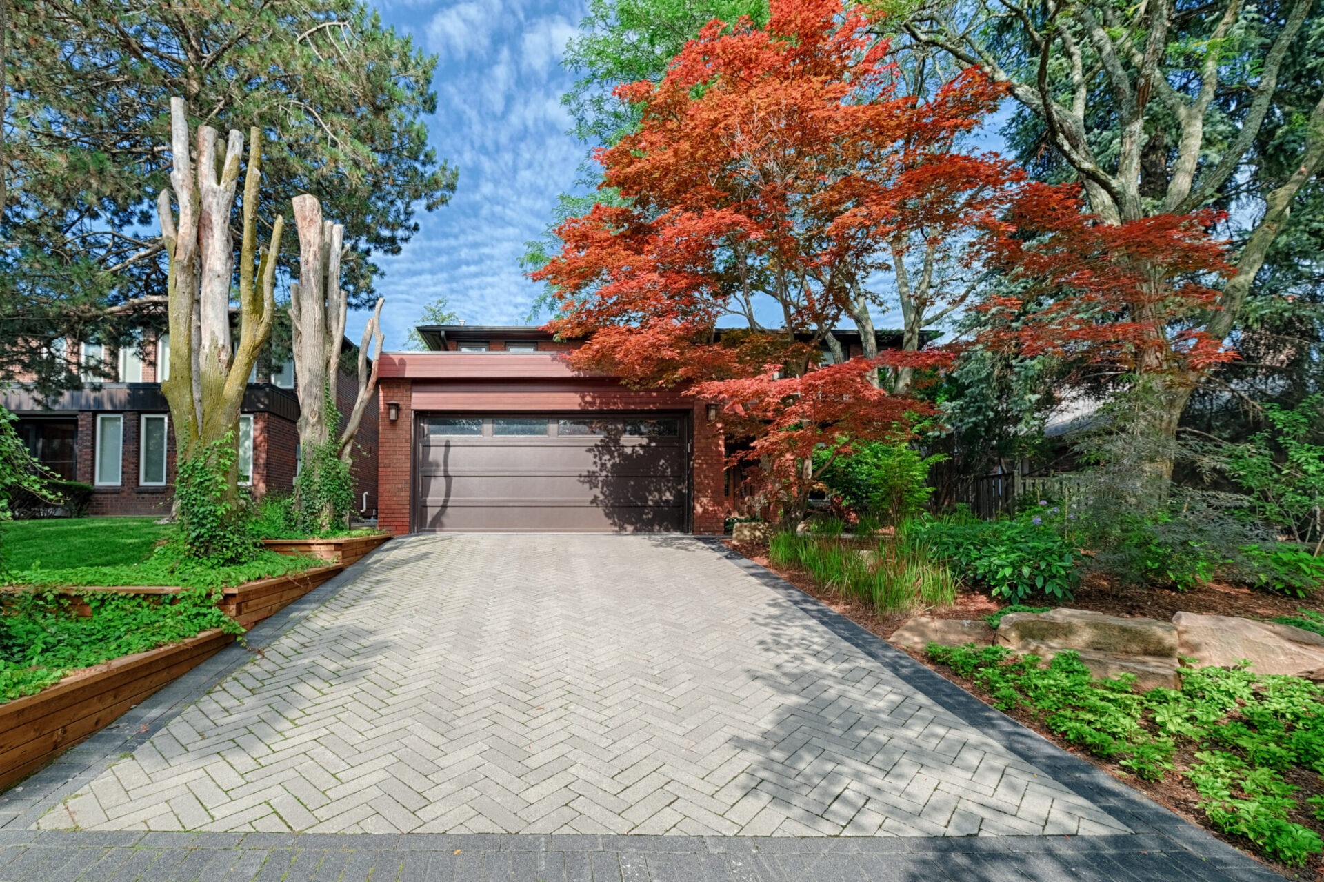 This image shows a residential driveway leading to a garage with a vibrant red tree on the right and pruned trees on the left, under a blue sky.