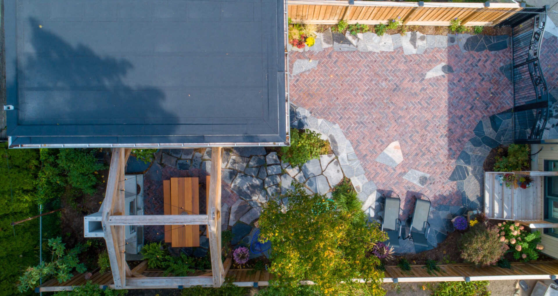 Aerial view of a landscaped backyard with a mix of herringbone brick and large stone tiles, wooden structures, garden beds, and outdoor furniture.