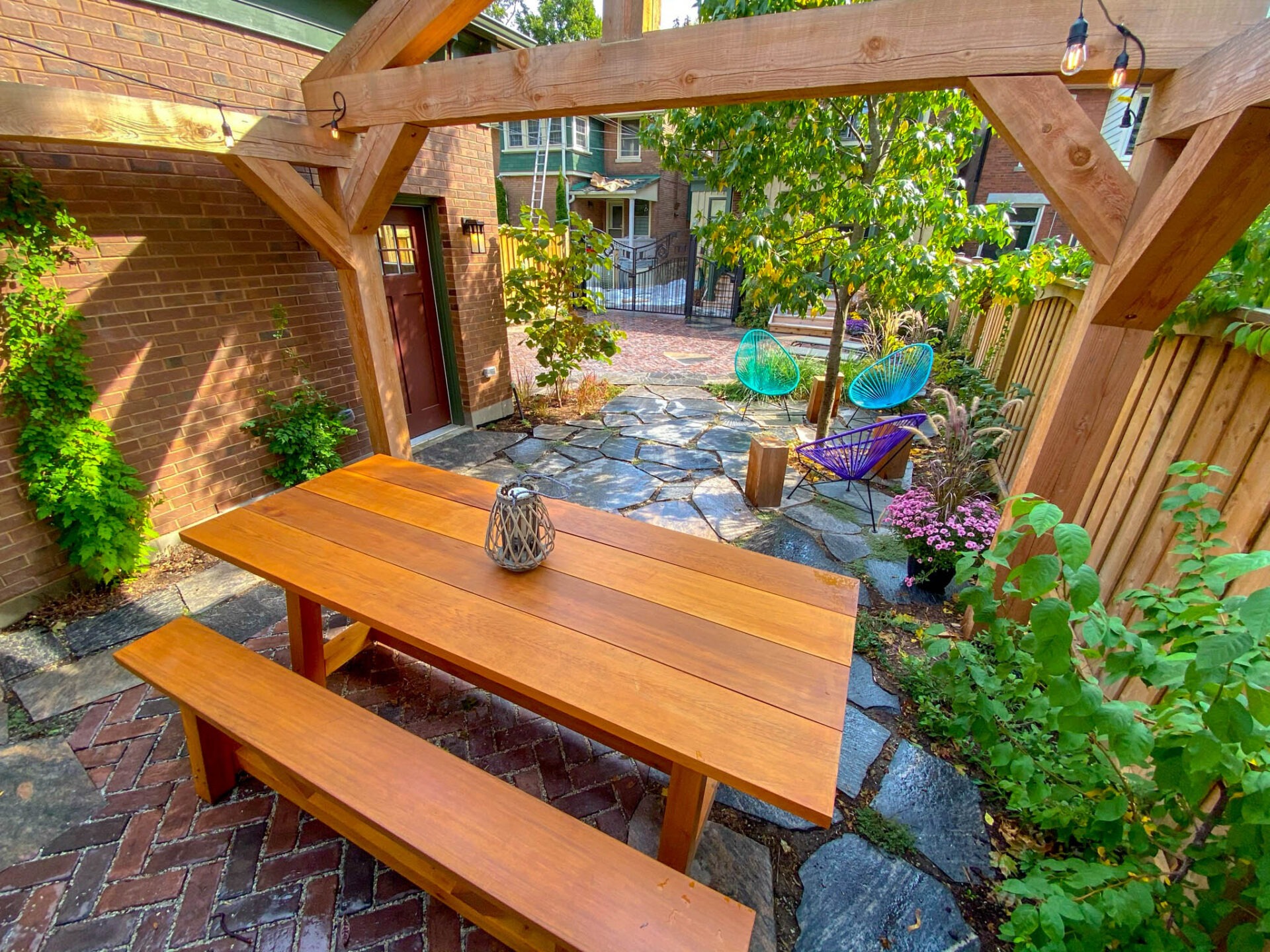 A cozy outdoor patio area with a wooden dining table and benches, string lights, brick floor, colorful chairs, plants, and a timber pergola.