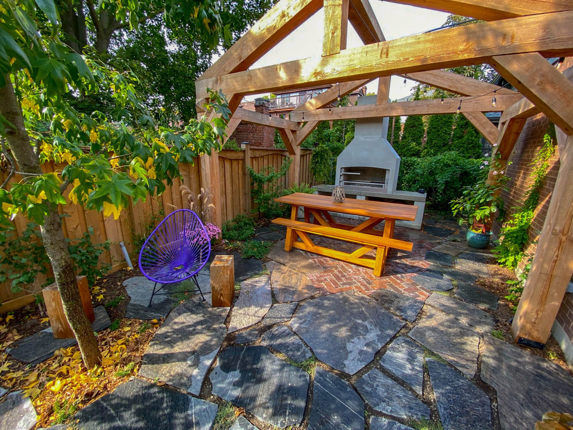 A serene outdoor space featuring a wooden pergola, pizza oven, wooden bench, a decorative purple chair, stone paving, and lush greenery with wooden fencing.