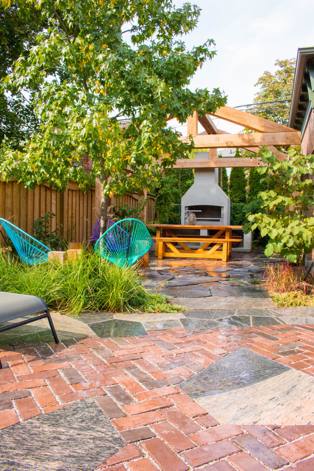 A cozy backyard with a brick and stone pathway, vibrant blue chairs, lush greenery, and a wooden pergola over an outdoor fireplace.