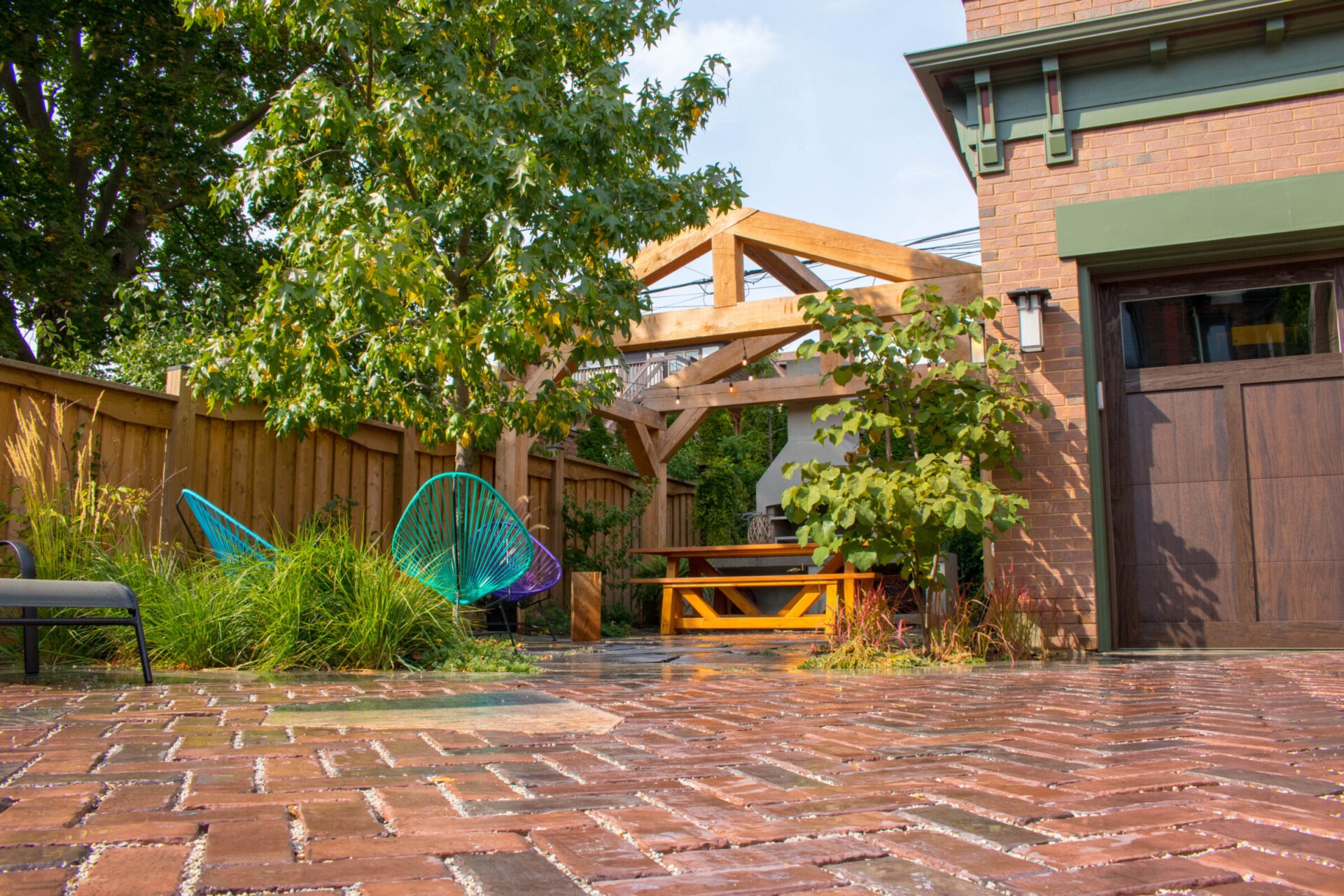 A brick patio with colorful chairs, wooden pergola, greenery, and part of a brown house on a sunny day. It's a peaceful, private outdoor space.