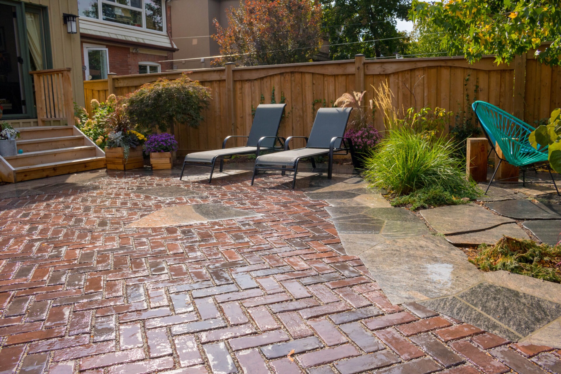 A sunny backyard with a red brick patio, two lounge chairs, wooden steps, vibrant plants, and a blue Acapulco chair by a wooden fence.