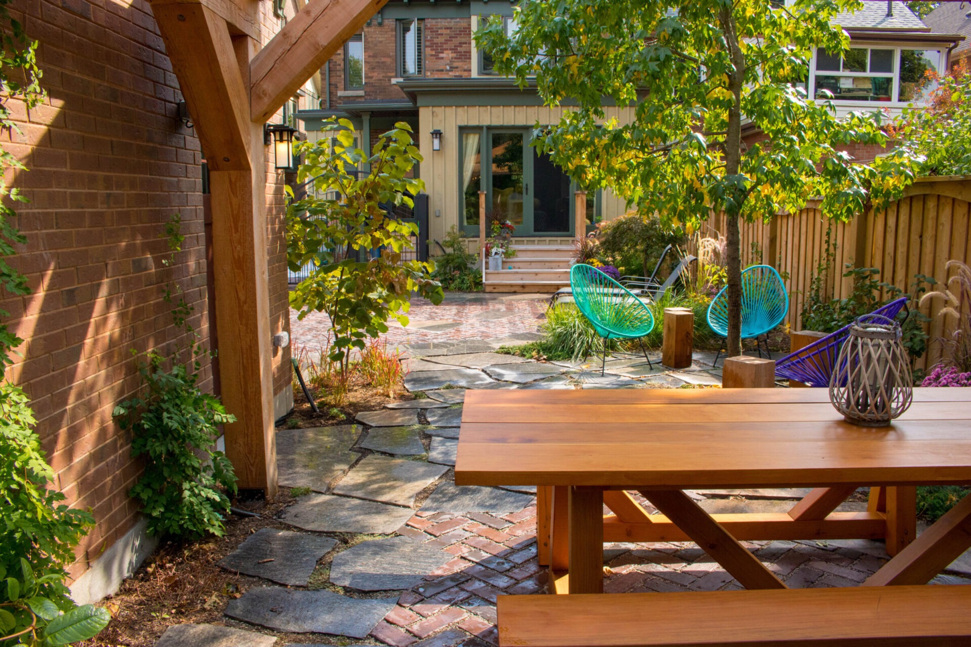 A serene backyard with stone pathways, a wooden picnic table, and vibrant blue-green wire chairs. Lush greenery surrounds a cozy, inviting outdoor space.