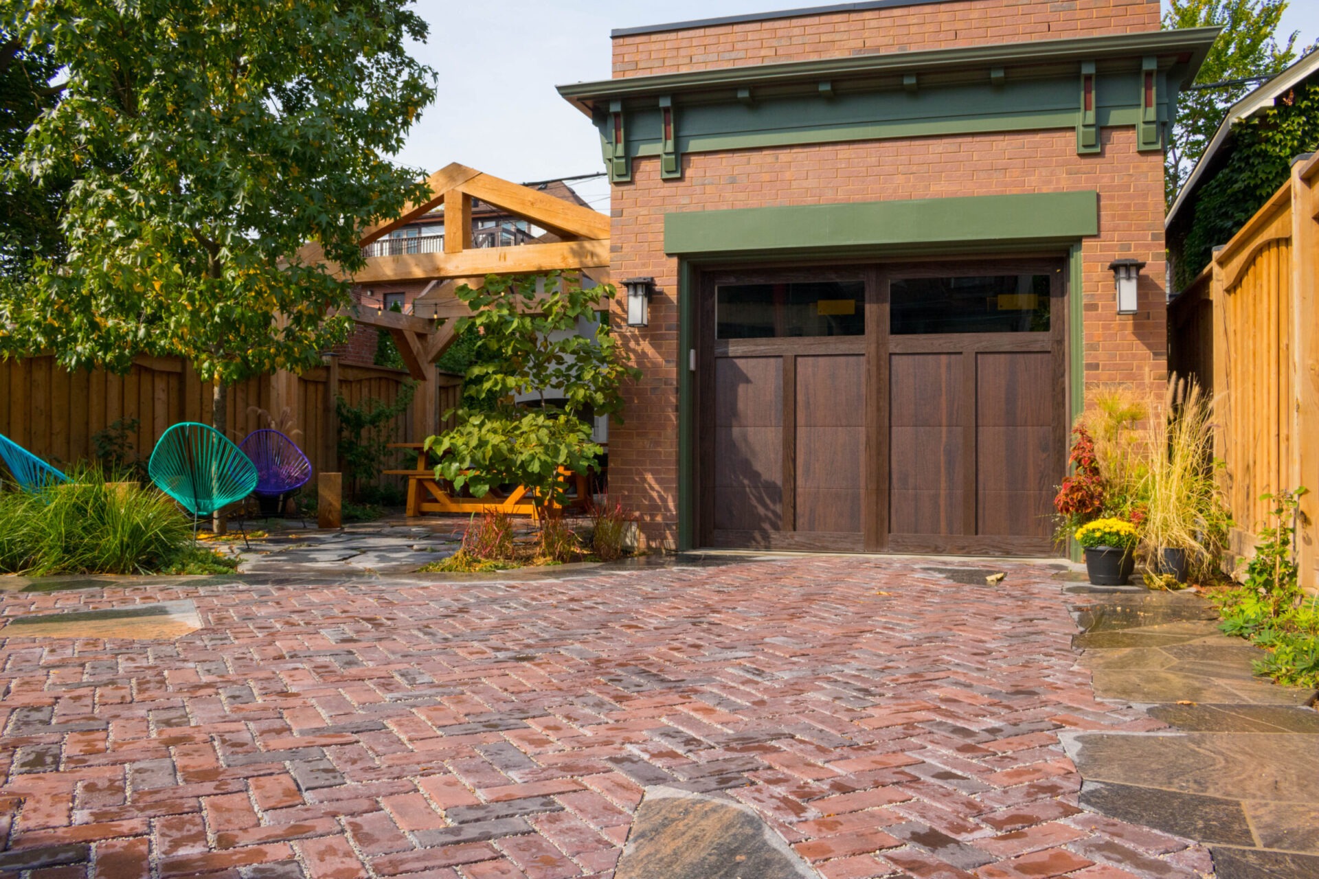 A red brick driveway leads to a garage door in a residential area, with a wooden pergola and colorful Acapulco chairs in a lush garden.