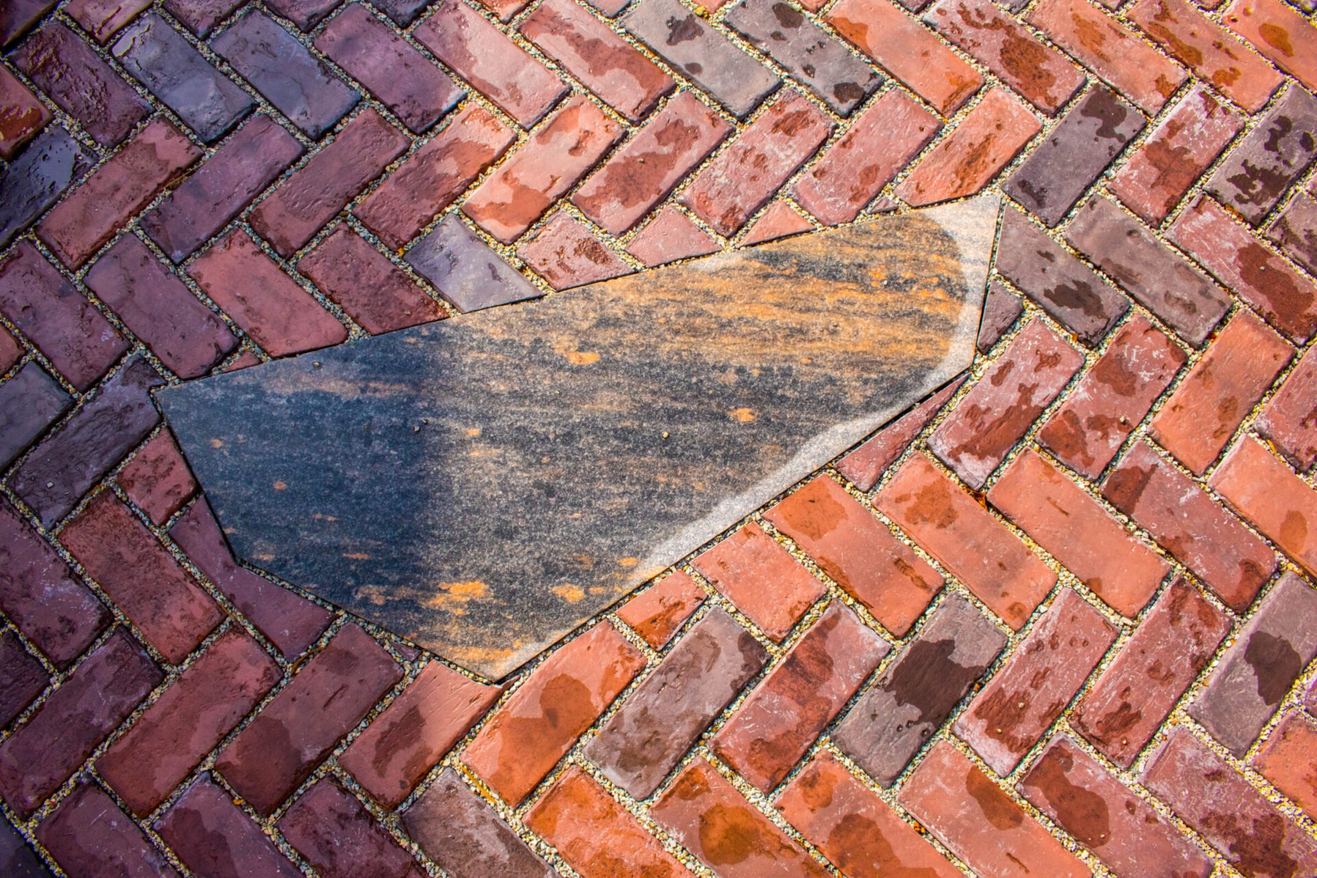 This image shows a wet, herringbone-patterned brick pavement with a large, irregular-shaped worn stone inset amongst the bricks.