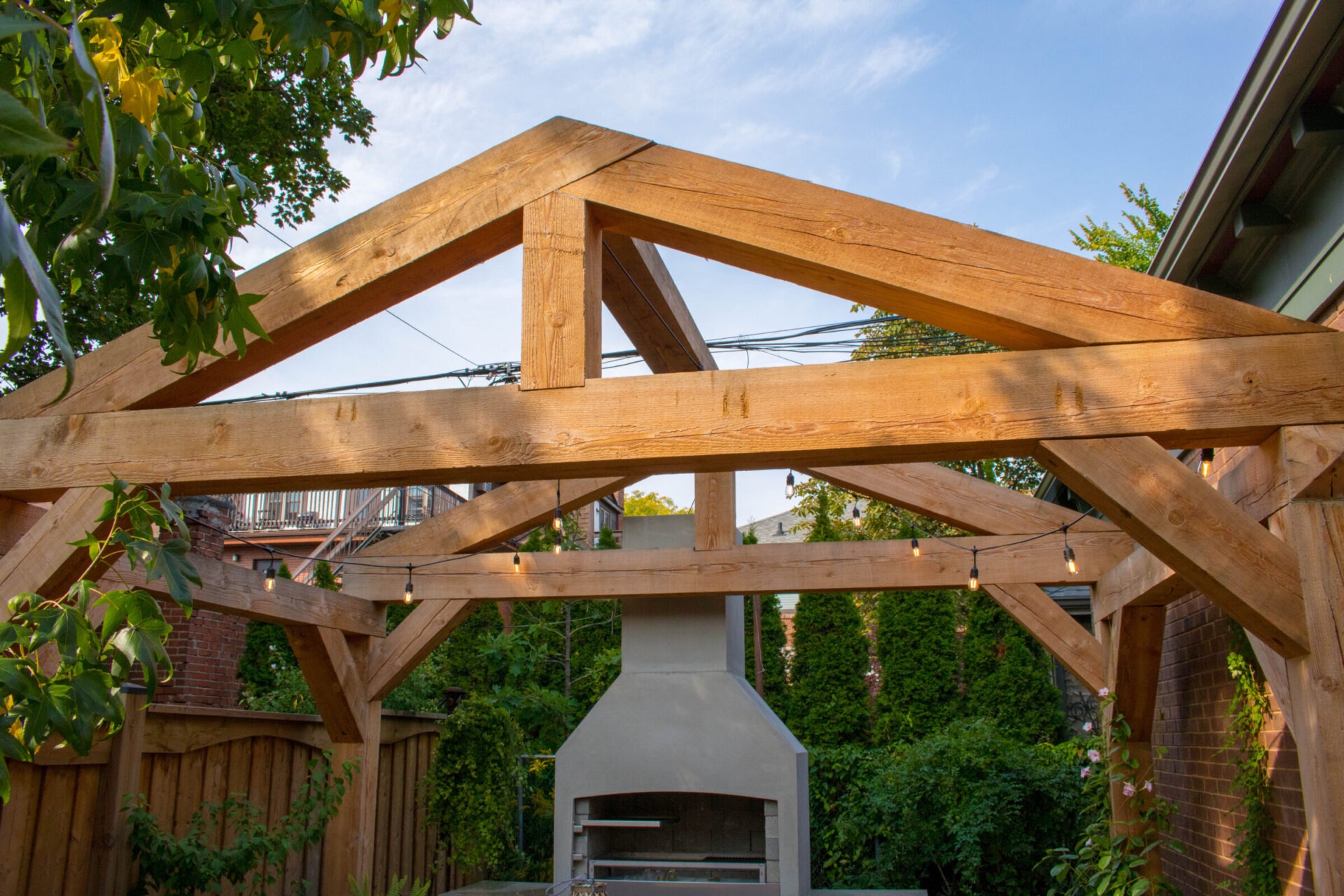 A wooden pergola structure in a backyard with string lights, a clear sky above, and a concrete outdoor fireplace in the background.